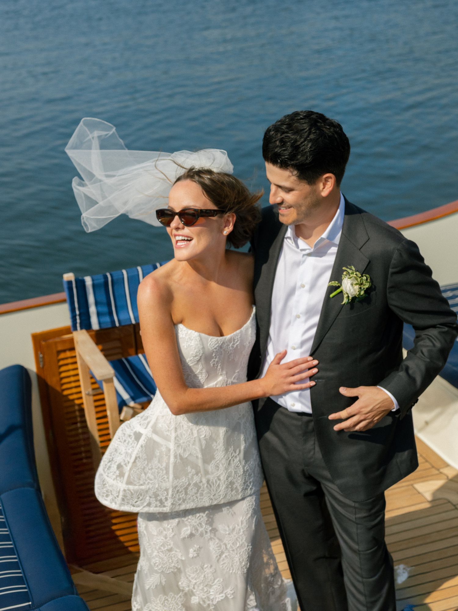 Smiling bride in lace dress with windswept veil leans on groom in dark suit on yacht deck.