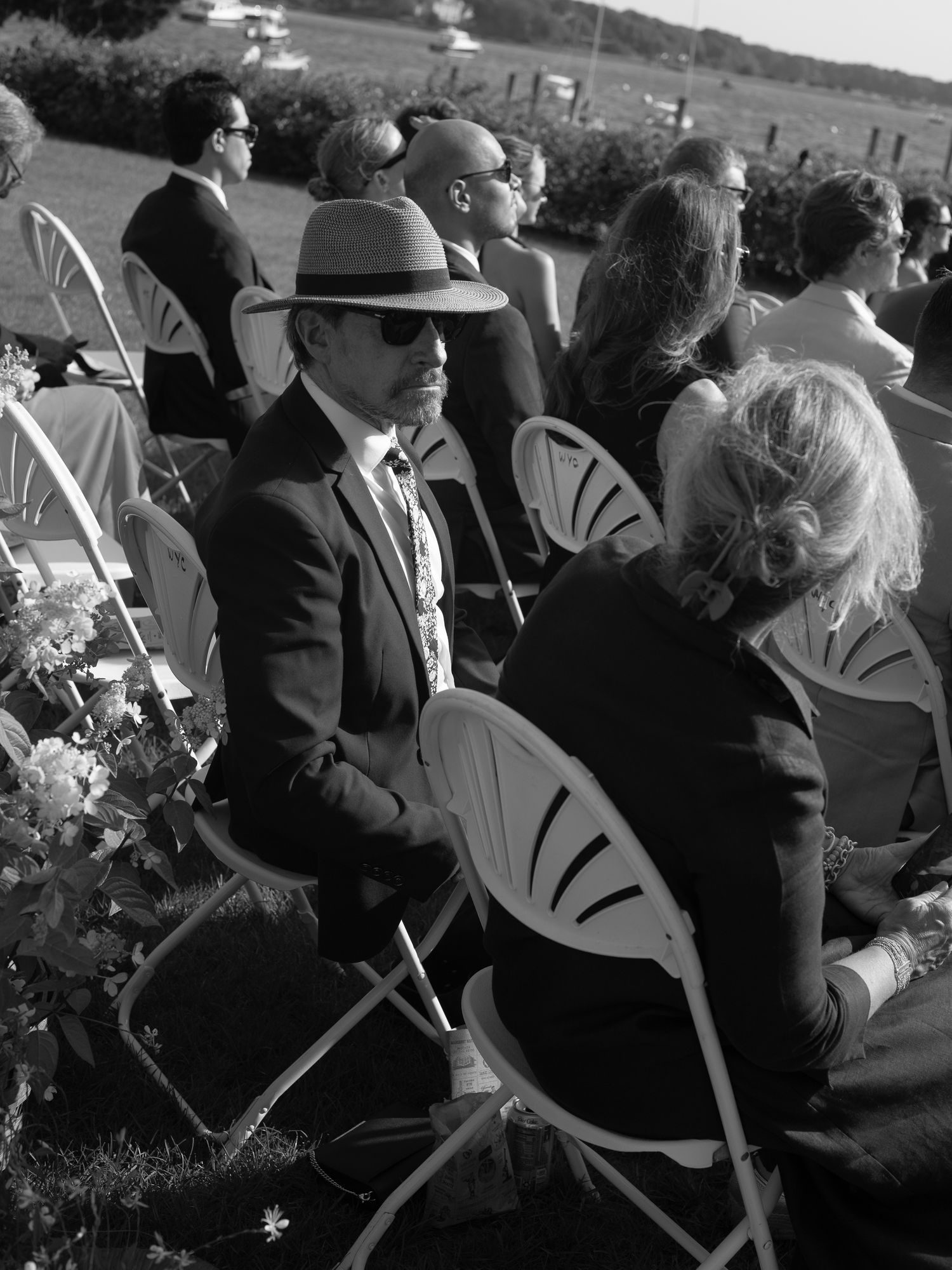 Black and white photo of wedding guests seated outdoors, man in hat and sunglasses in foreground.