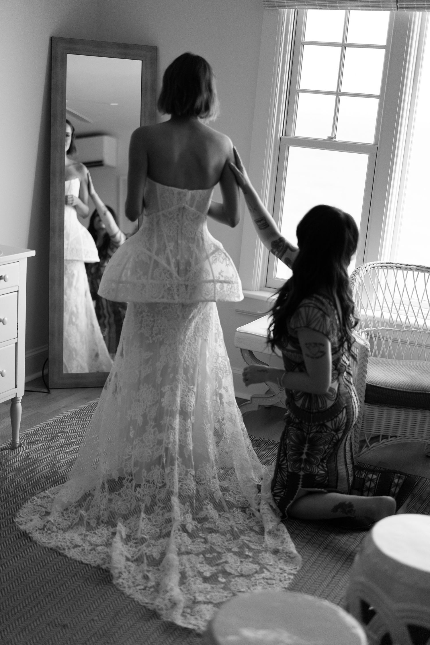 Black and white photo of bride in lace peplum gown being helped by attendant before wedding.