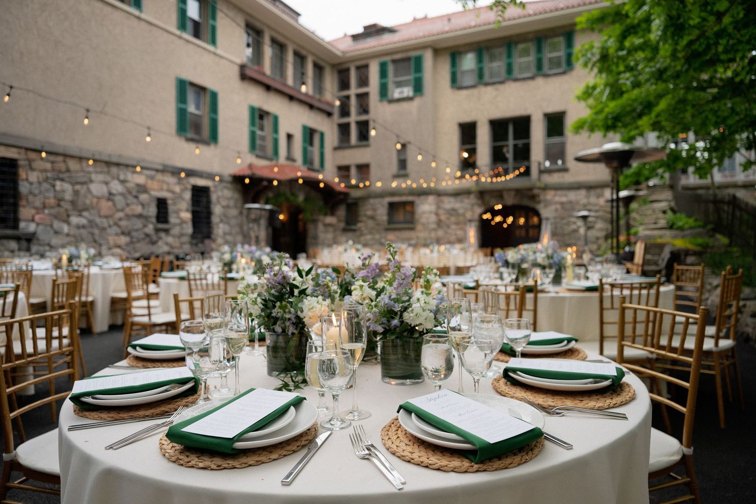 Elegant outdoor wedding reception courtyard with round tables, gold chiavari chairs, and string lights.