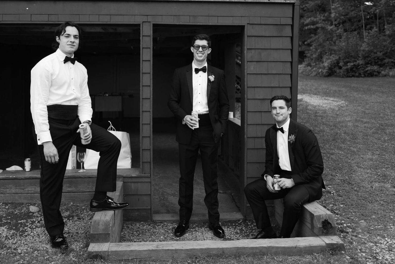 Three groomsmen in tuxedos pose outside a rustic cabin in black and white photo.