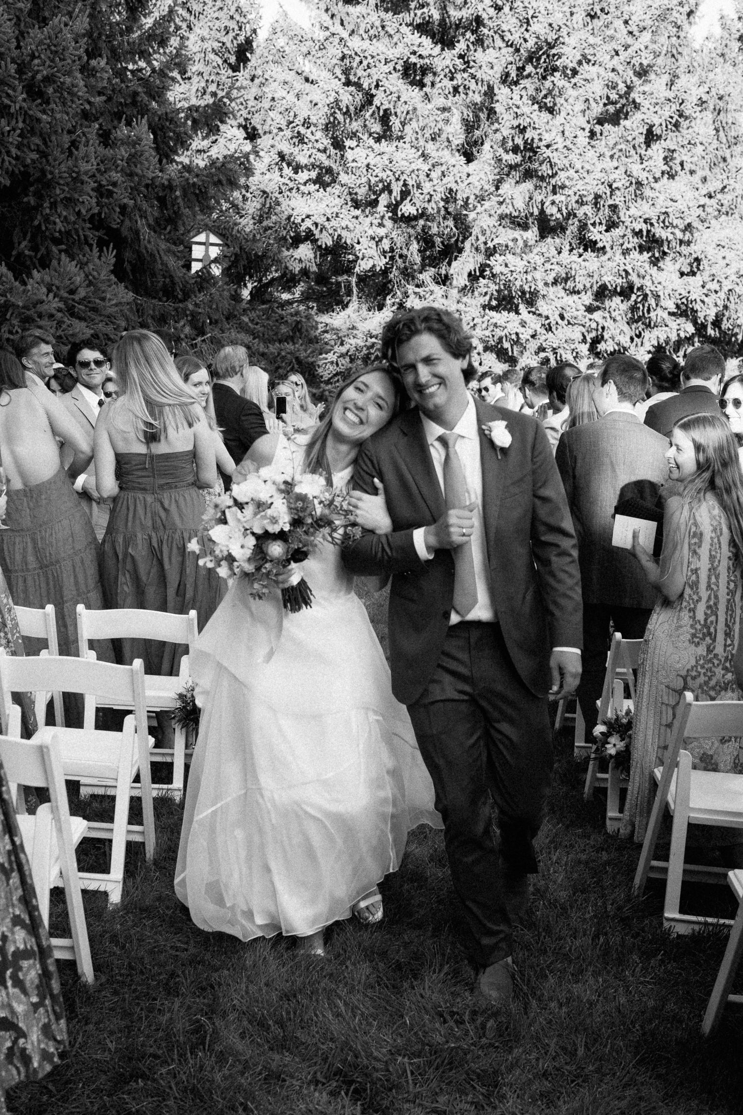 Black and white photo of joyful newlyweds walking back down the aisle after outdoor wedding ceremony.