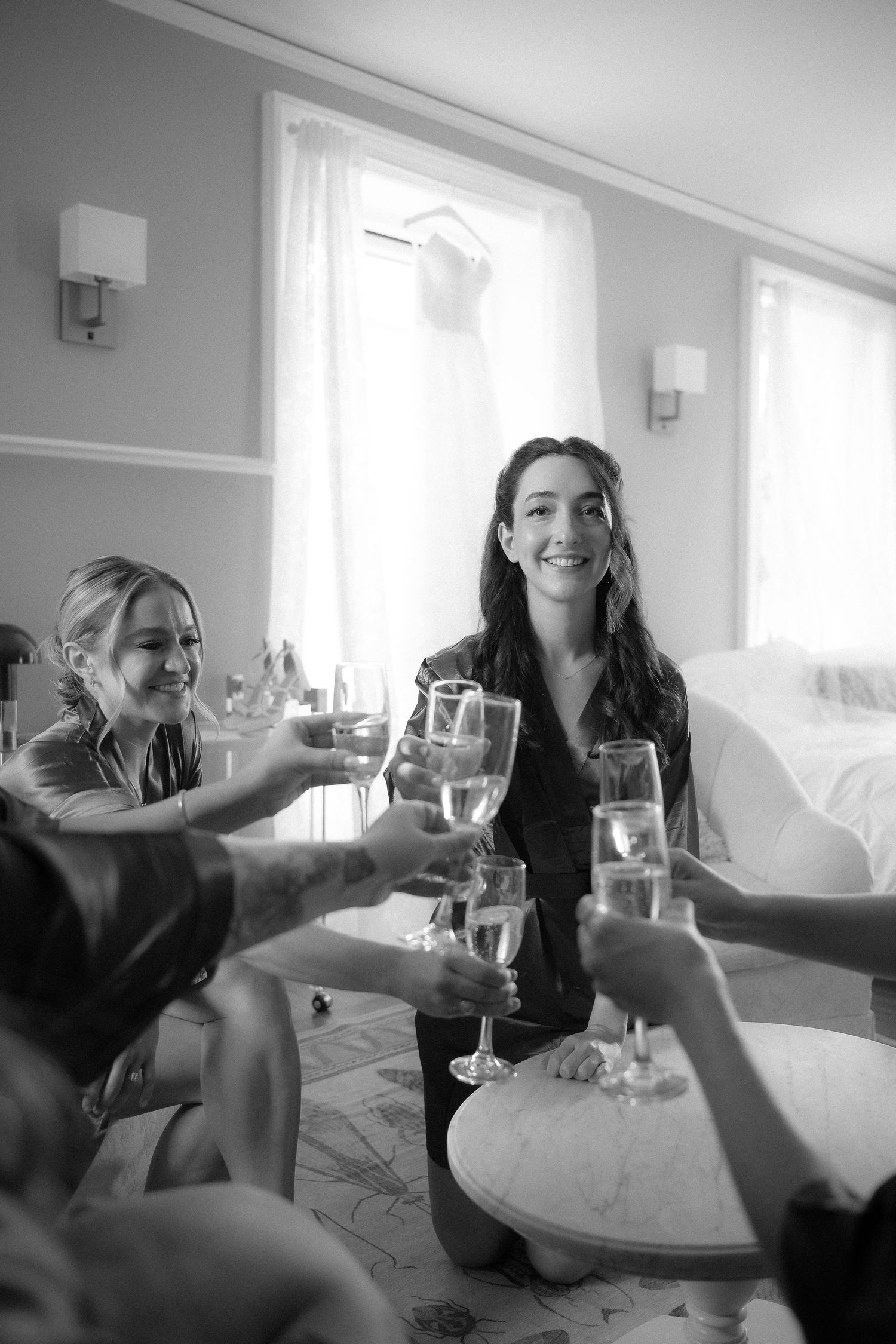 Bridesmaids in silk robes toasting with champagne glasses in a bright hotel room before the wedding.
