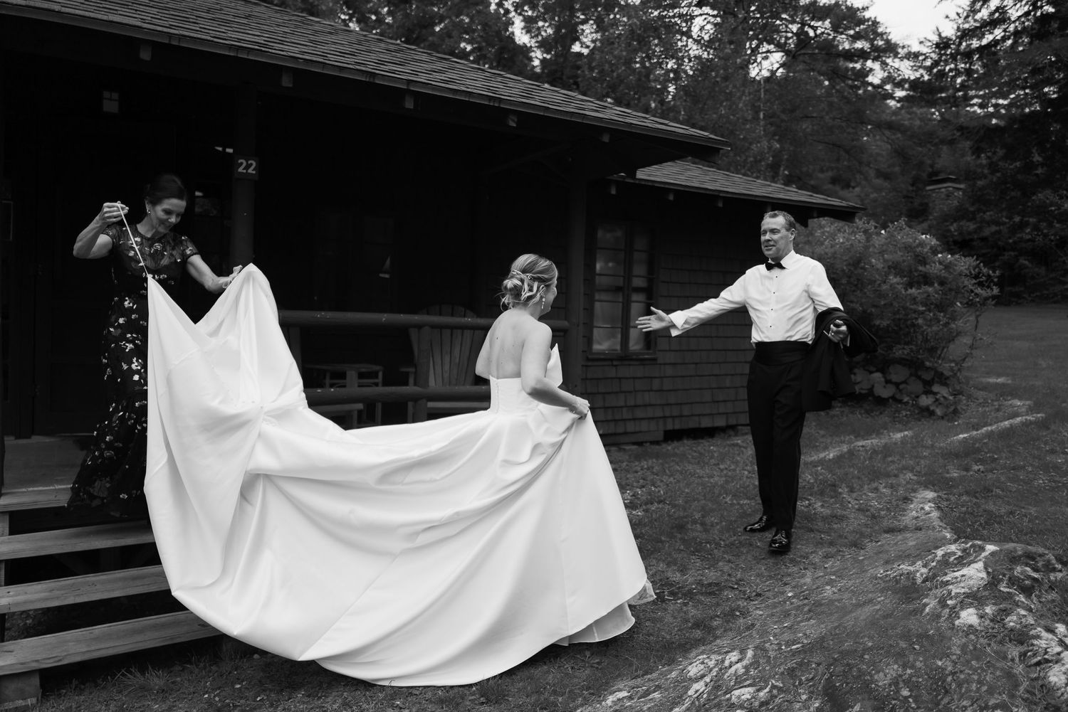 Bridesmaids help bride with wedding gown train outside rustic cabin in black and white.