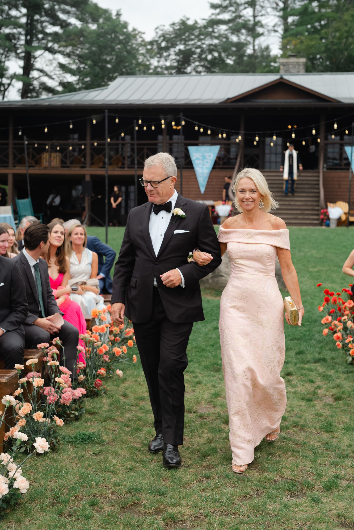 Elegant couple walking down outdoor wedding aisle, man in tuxedo and woman in blush off-shoulder gown.