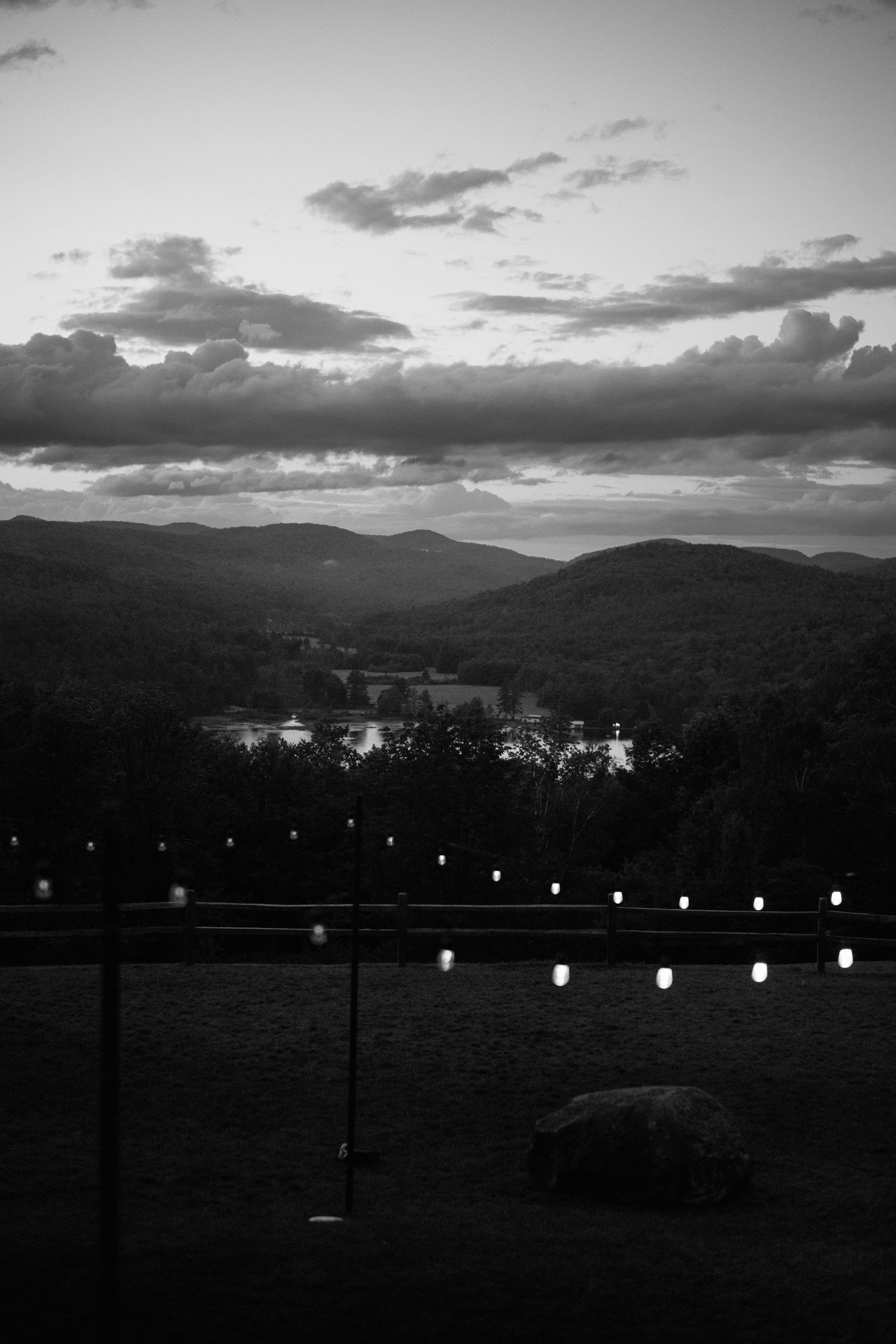 Black and white landscape of rolling mountains at sunset with string lights along a fence line.