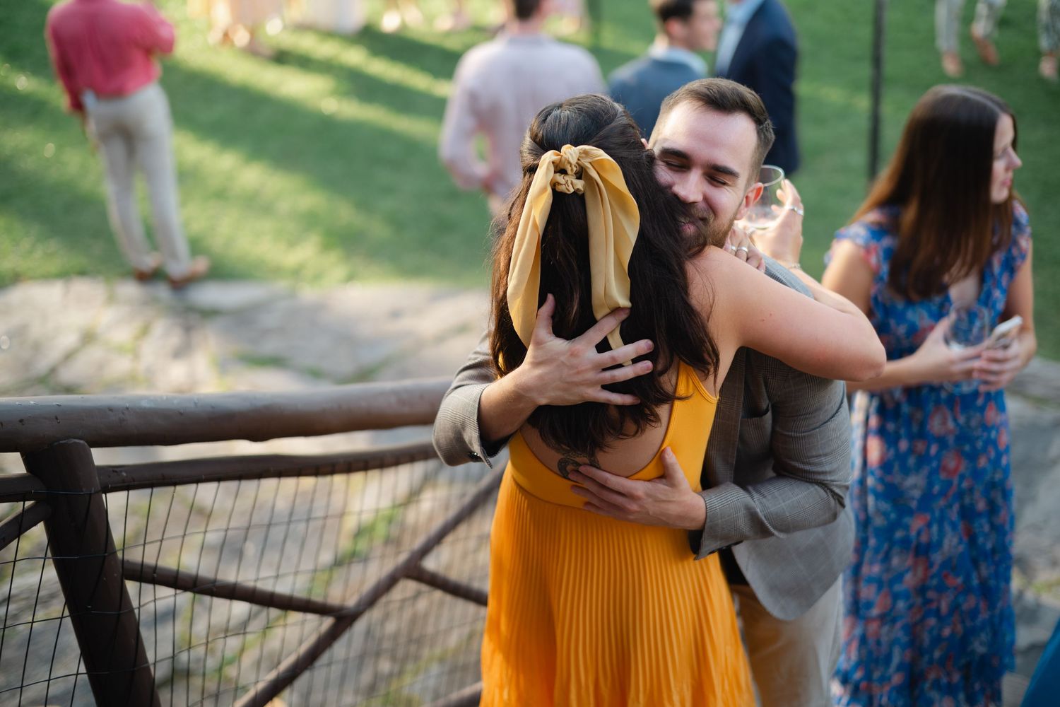 Wedding guest in yellow dress receiving a warm hug from a man holding a wine glass outdoors.