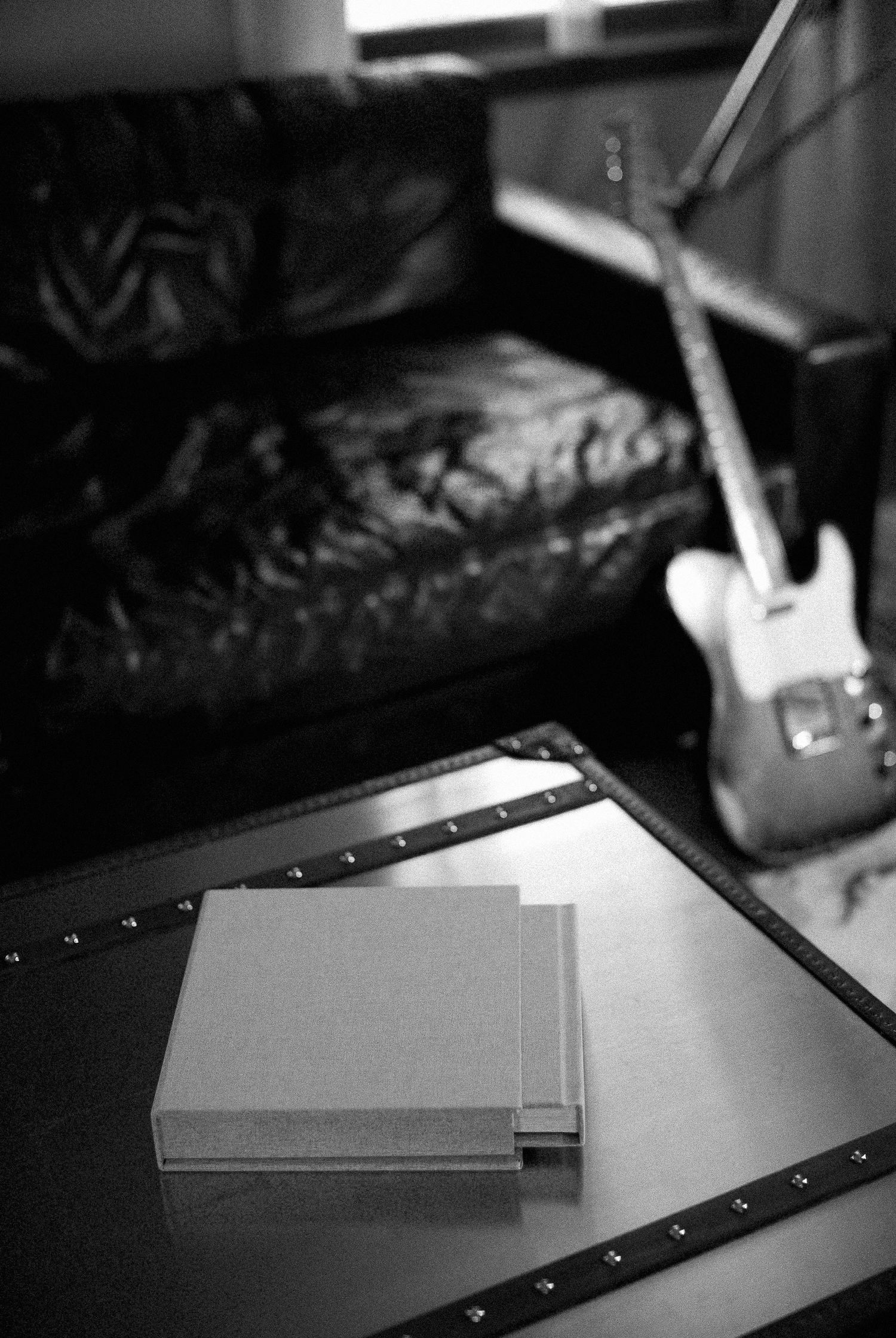 Monochrome photo of books stacked on metal trunk table with leather sofa and electric guitar behind.