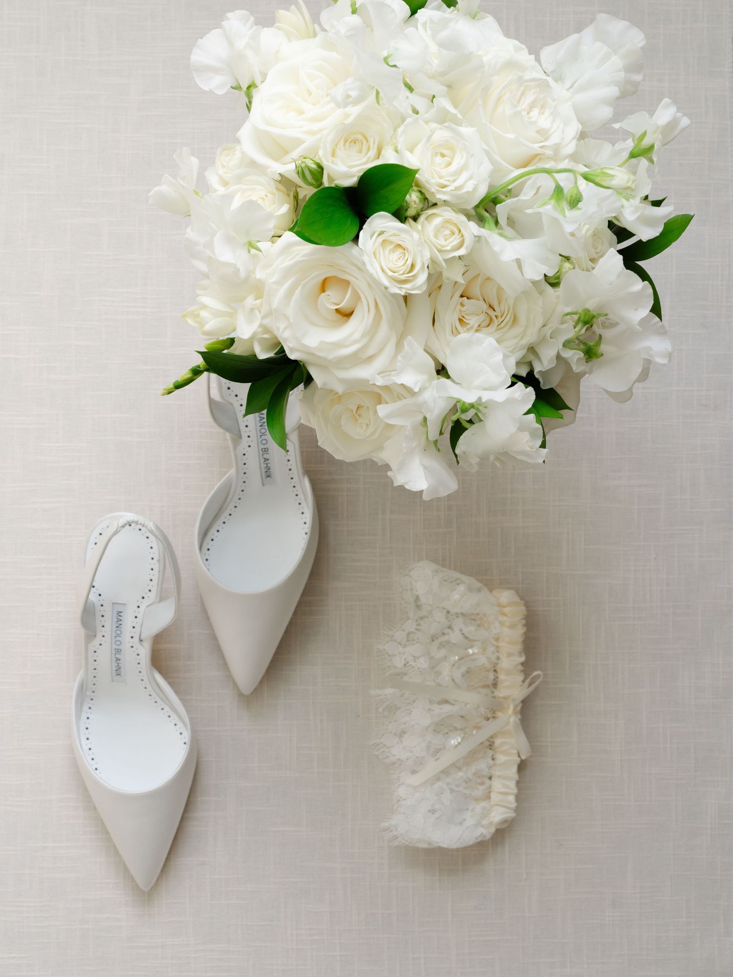 Elegant bridal flat shoes, white rose bouquet, and lace garter arranged on neutral linen background.