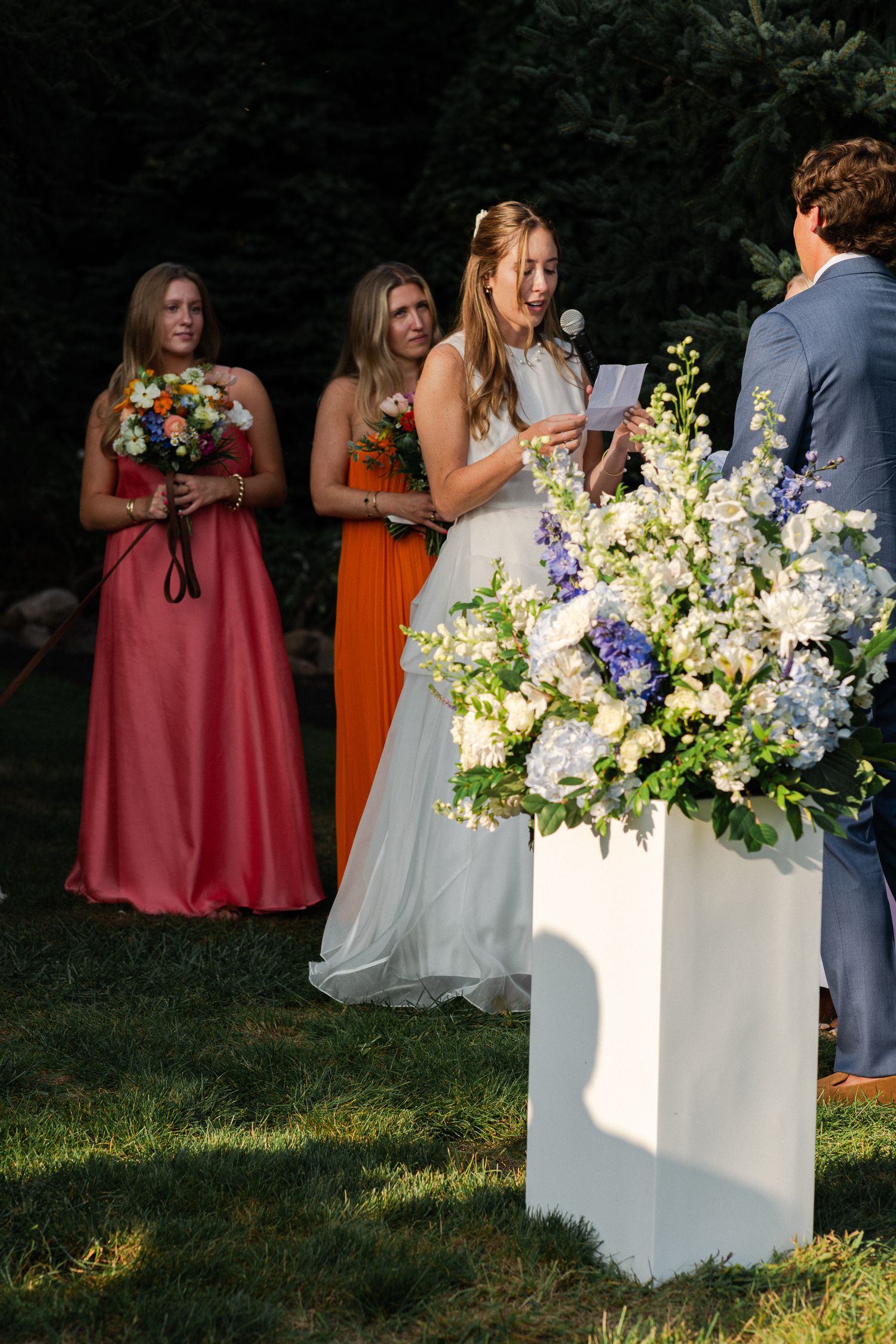 Bride reading heartfelt vows at outdoor wedding ceremony beside white floral arrangement with bridesmaids behind her.