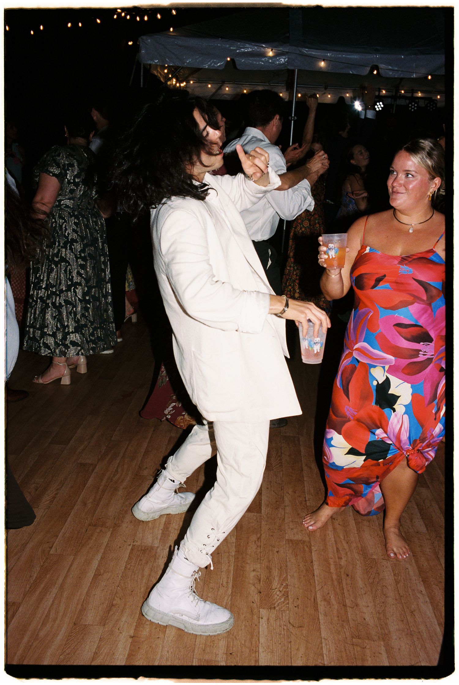 A guest in a white suit dances energetically at an outdoor wedding reception under string lights.