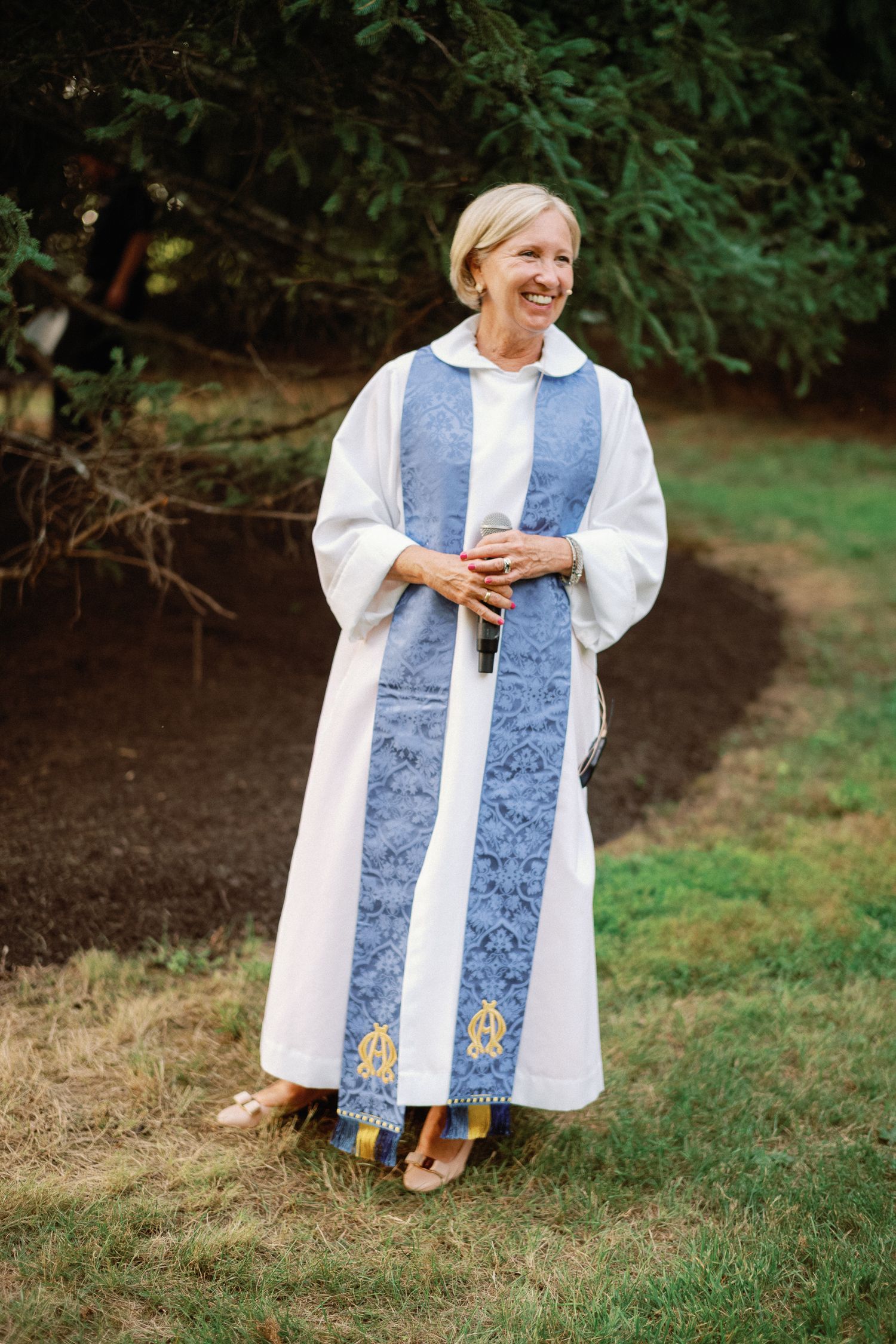 Smiling female officiant in white and blue ceremonial robes holding a microphone at an outdoor wedding.