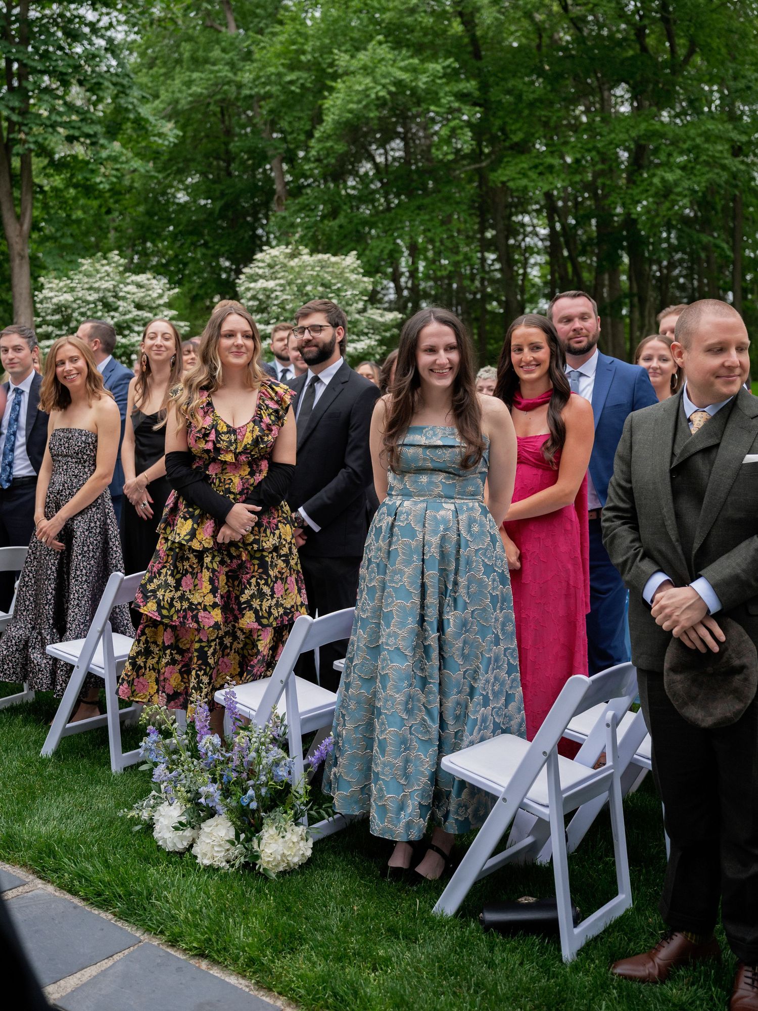 Wedding guests in colorful formal attire stand and watch an outdoor garden wedding ceremony.