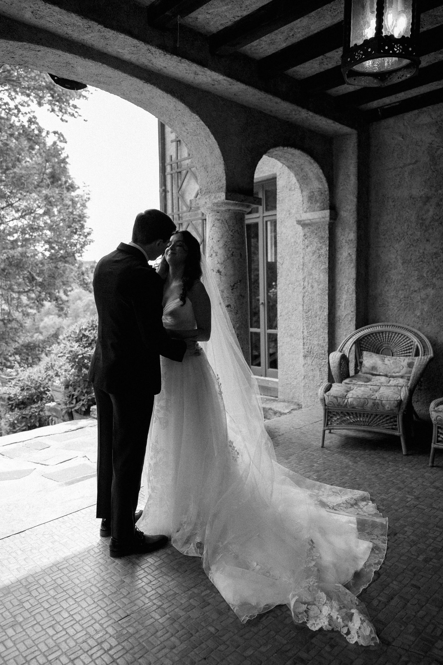Black and white photo of a bride and groom embracing under an arched stone portico.