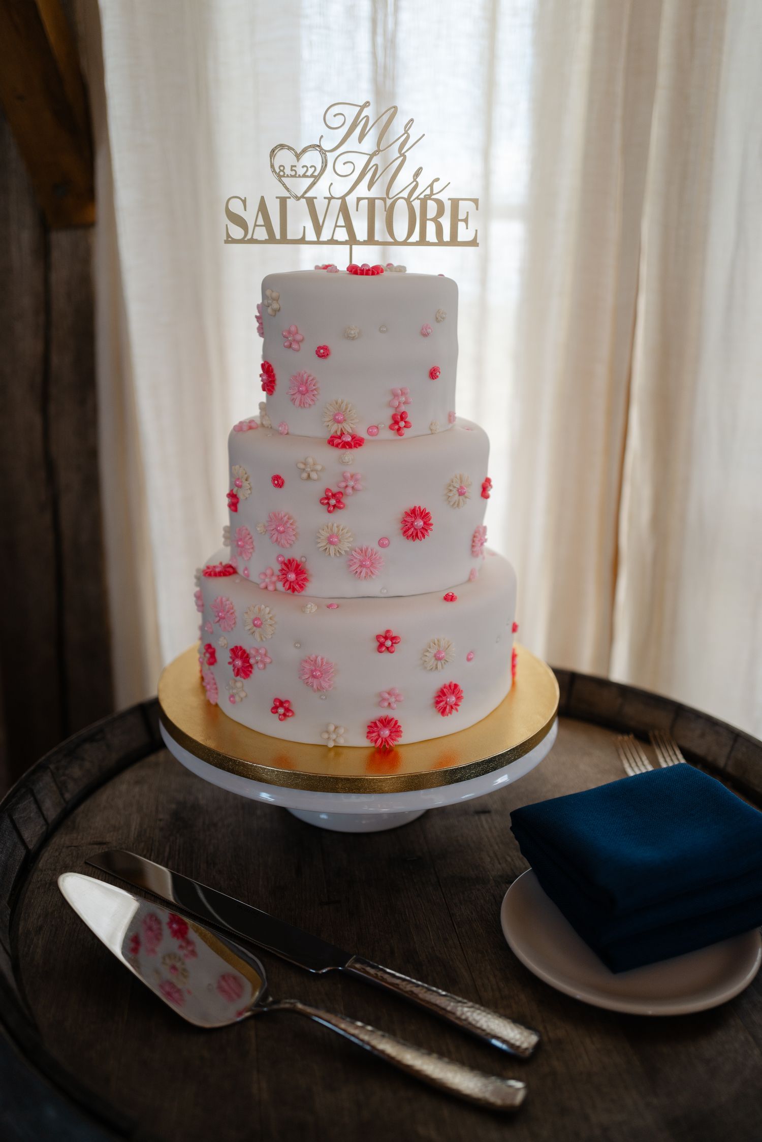 Three-tier white wedding cake with pink floral details and Mr. and Mrs. Salvatore gold cake topper.