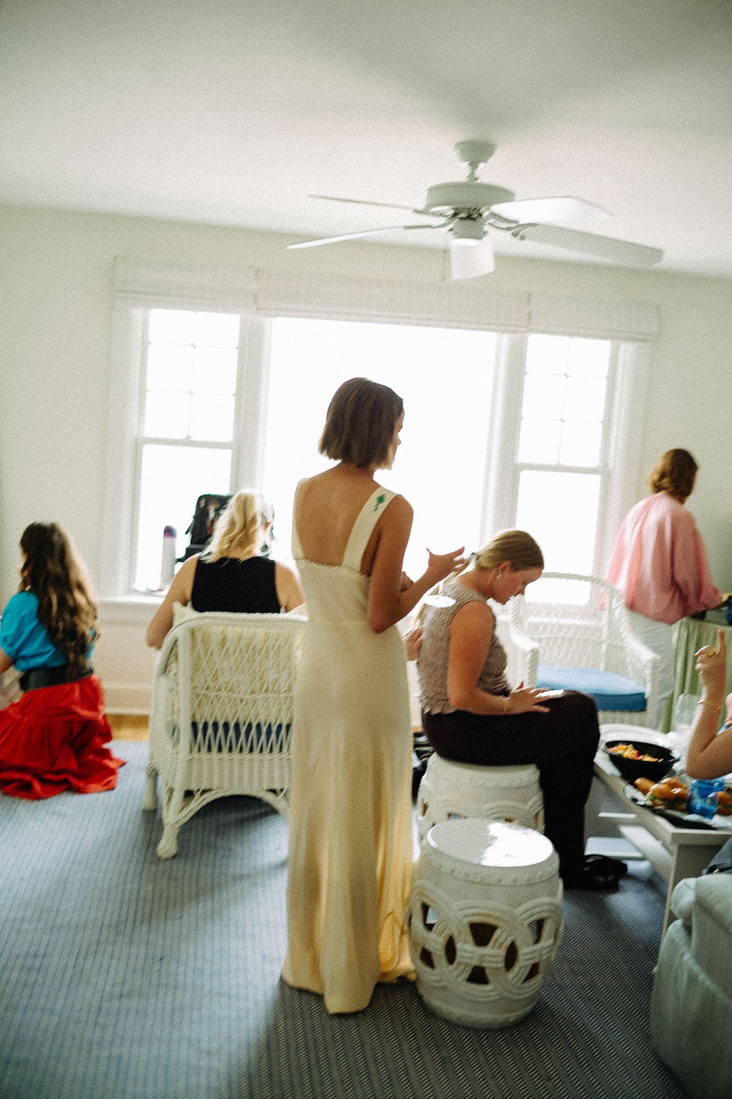 Bride in white backless gown surrounded by bridesmaids in bright room with ceiling fan during wedding preparation.