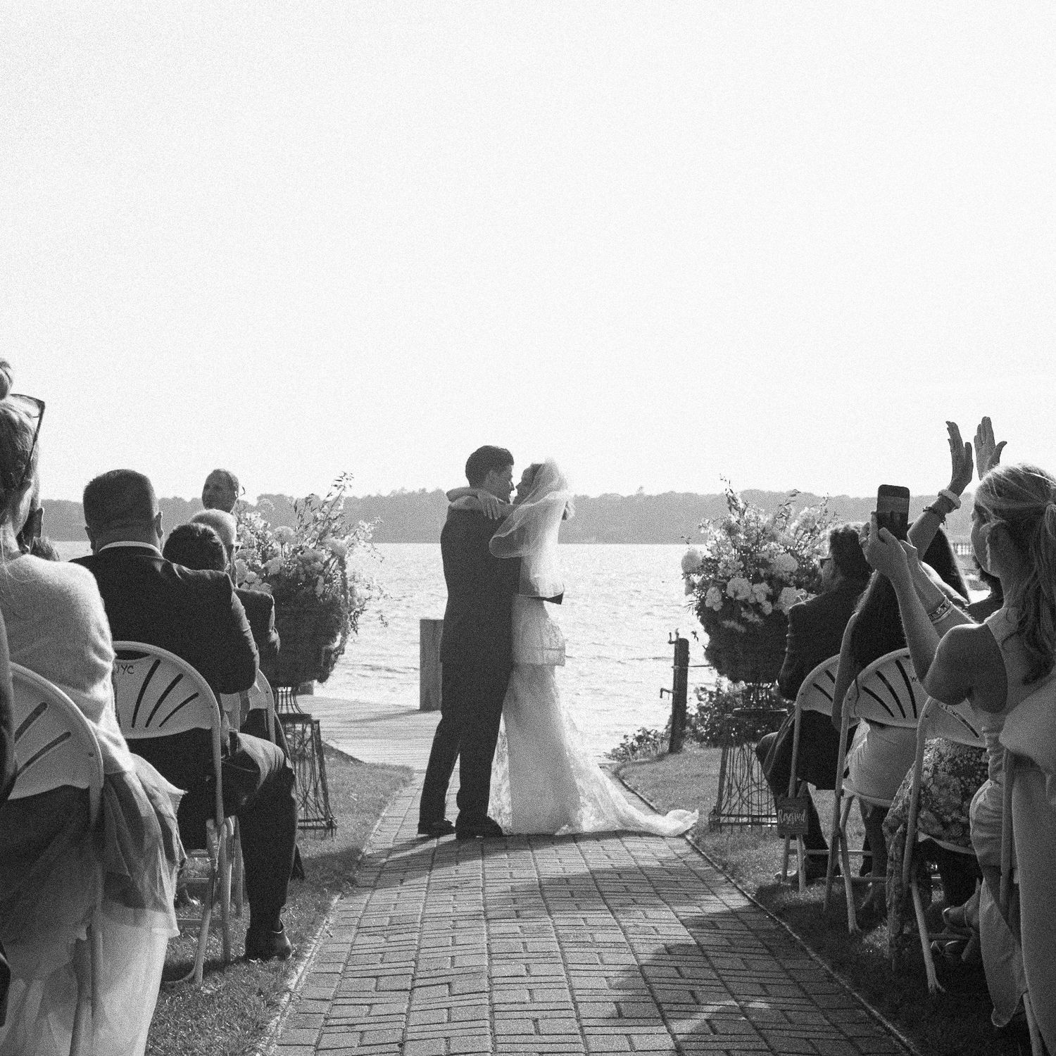 Black and white photo of a couple sharing their first kiss at an outdoor waterfront wedding ceremony with seated guests.