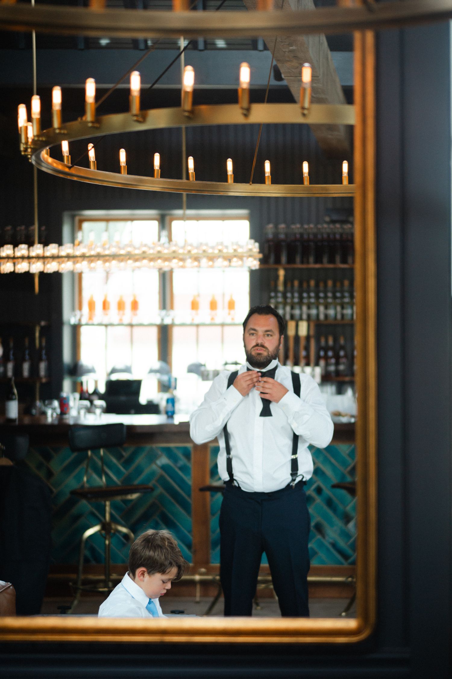 Groom adjusting bow tie reflected in gold mirror at elegant bar venue.