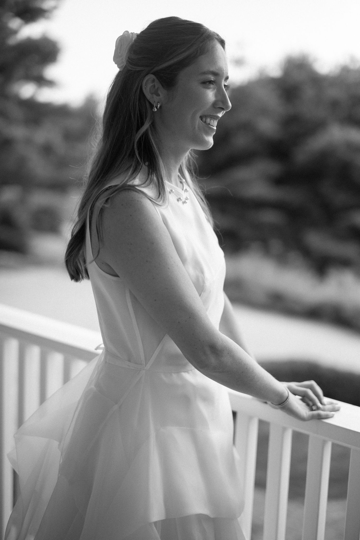 Black and white portrait of a smiling bride in a white dress leaning on a railing outdoors.