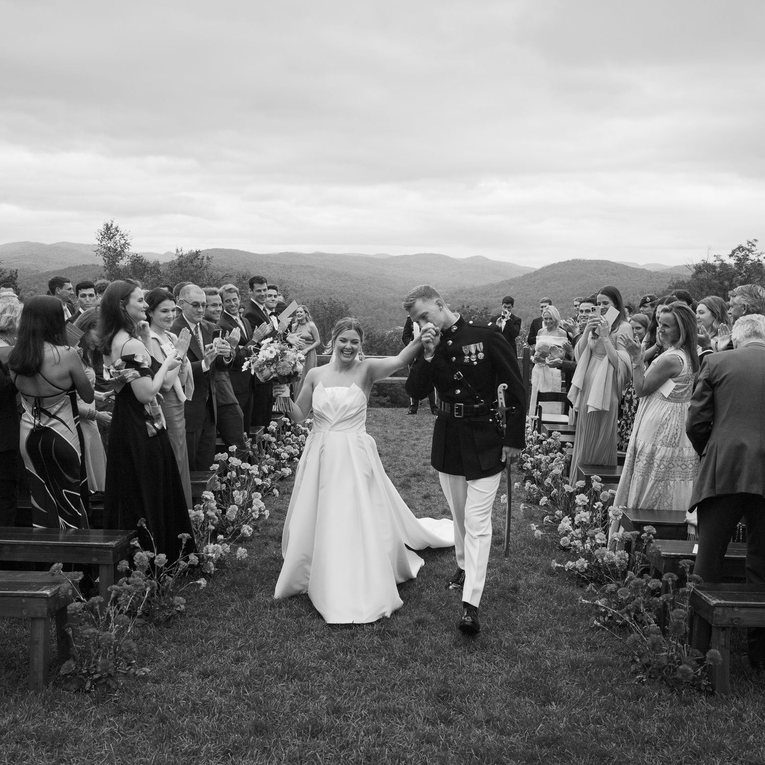 Black and white photo of groom in military uniform escorting bride down aisle as guests celebrate.