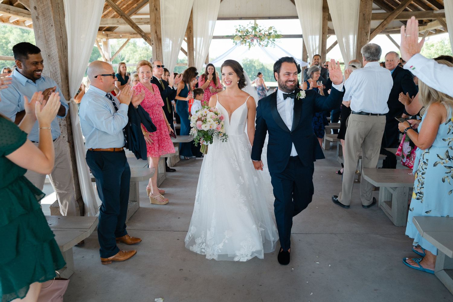 Bride and groom walk down the aisle as guests cheer at an outdoor wedding ceremony under a wooden pavilion.