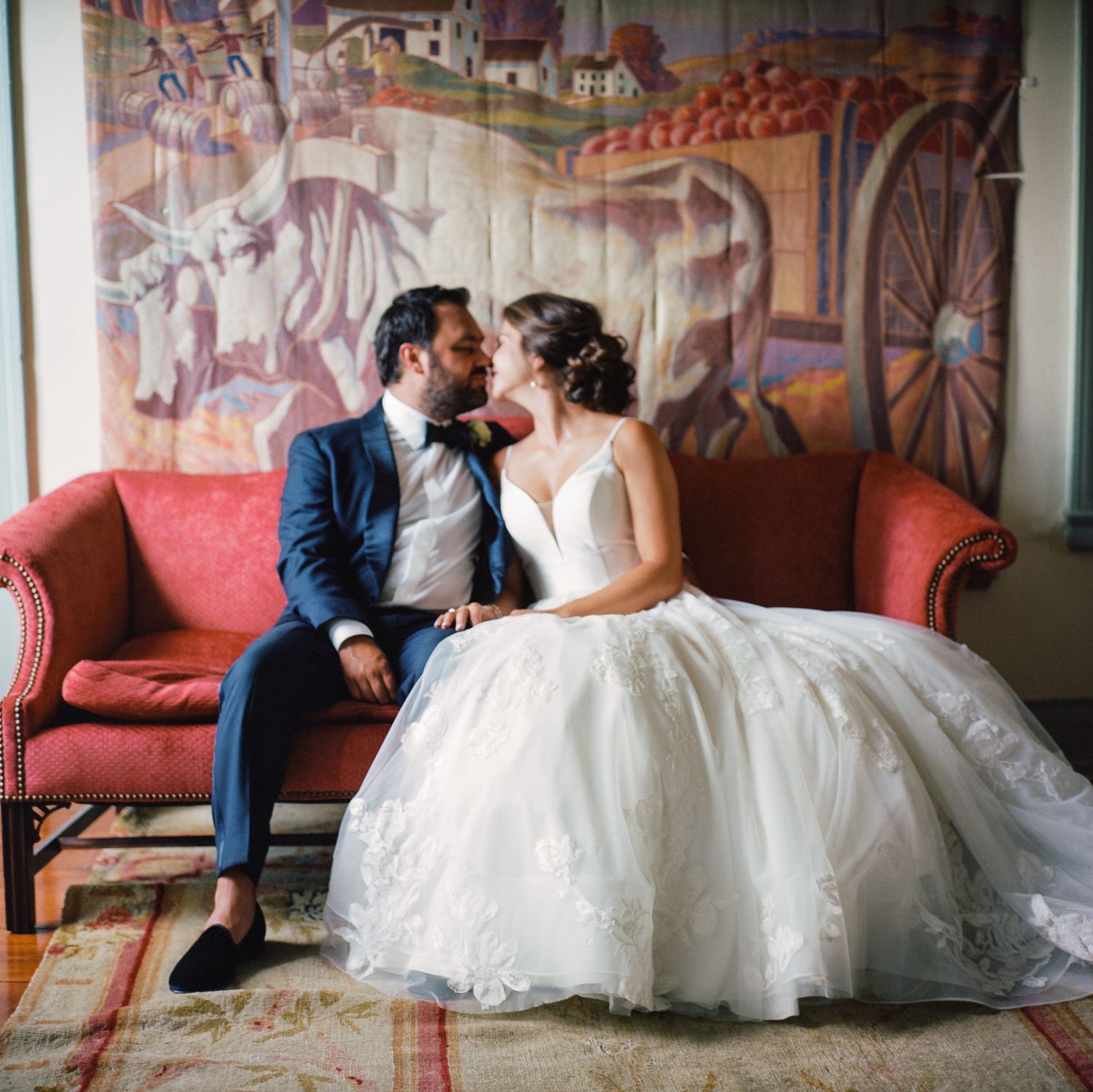 Bride in ball gown and groom in navy suit sharing kiss on red sofa before rustic mural.