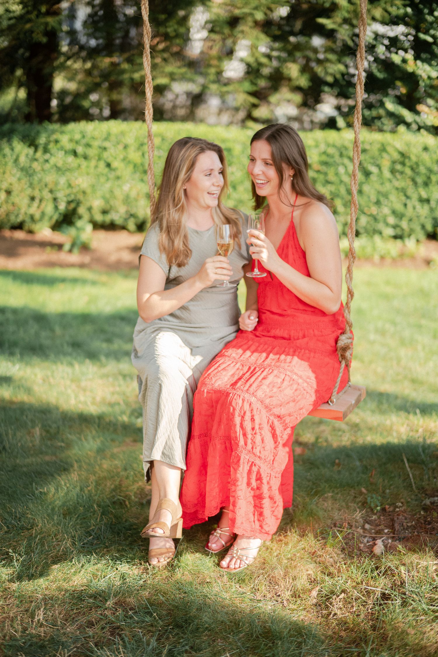 Two smiling women toasting with champagne glasses while sitting together on a rope swing outdoors.