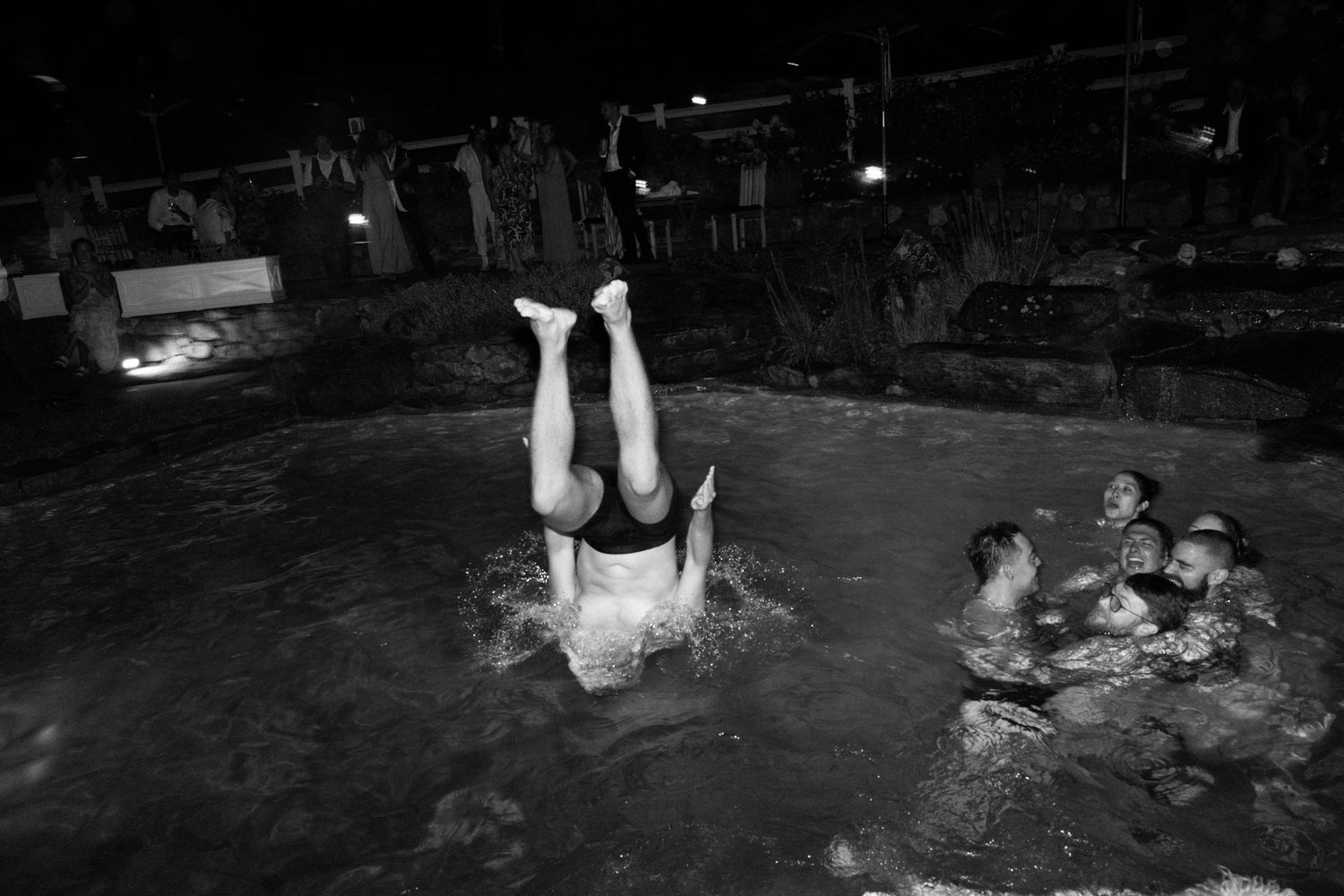 Wedding guest doing a handstand dive into pool surrounded by swimmers at nighttime wedding celebration.