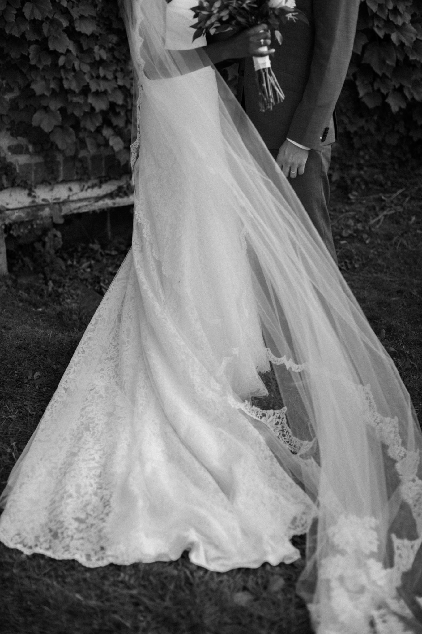 Black and white wedding photo showcasing bride's lace gown and flowing veil beside groom outdoors.