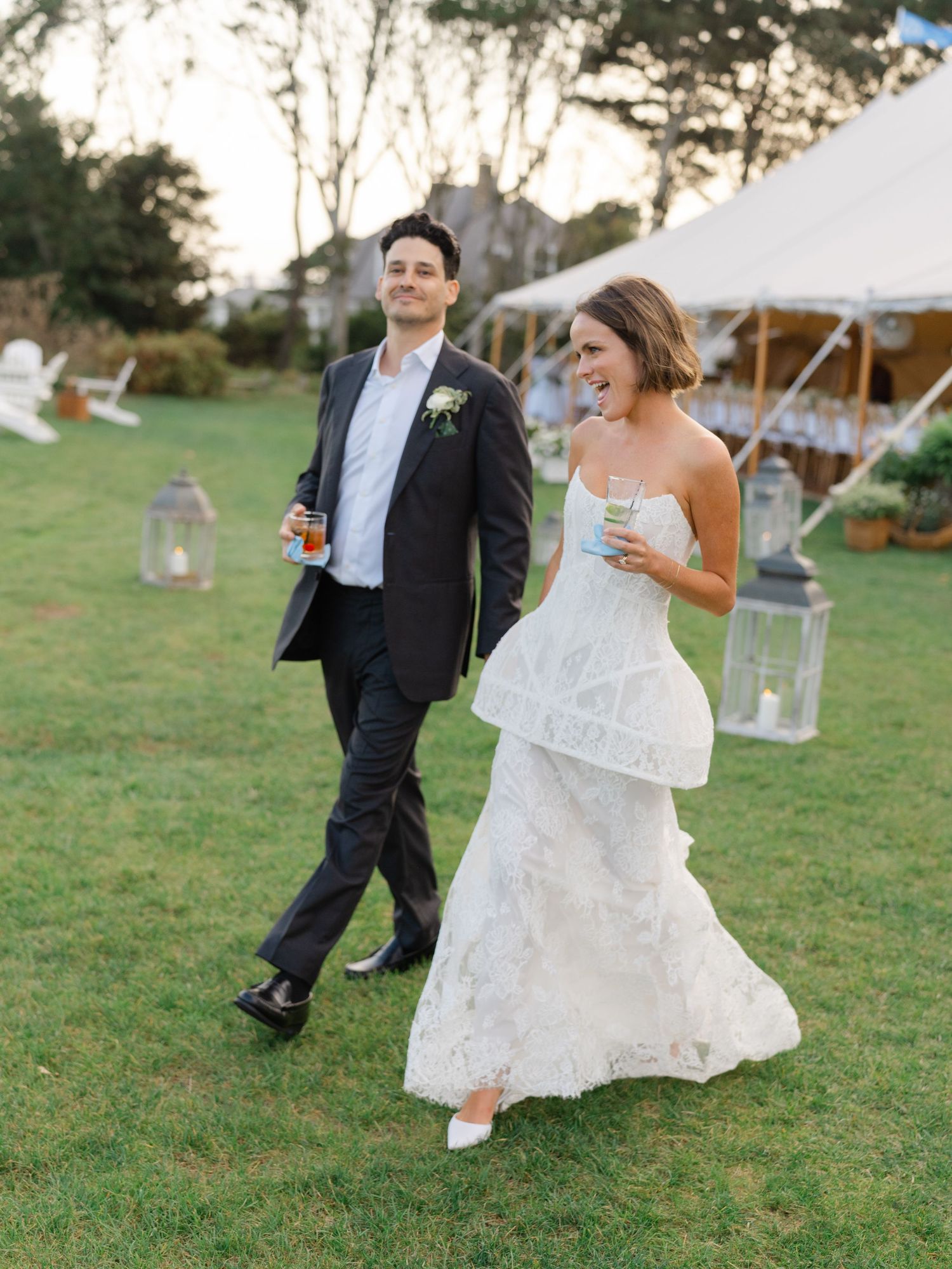 Bride and groom walk together on lawn at dusk holding drinks near a white wedding reception tent.