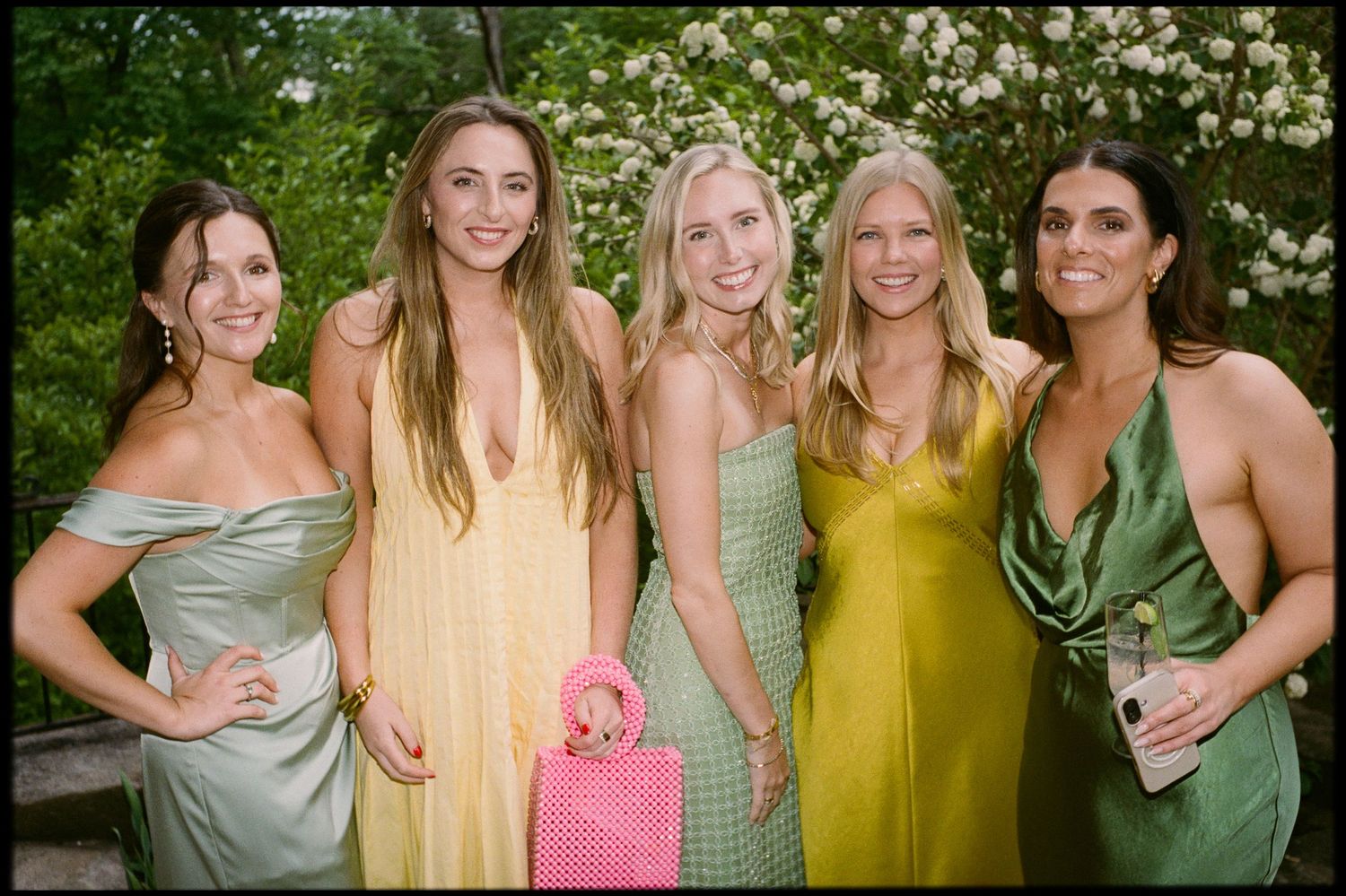 Five women in colorful formal gowns posing together outdoors at a spring wedding celebration.