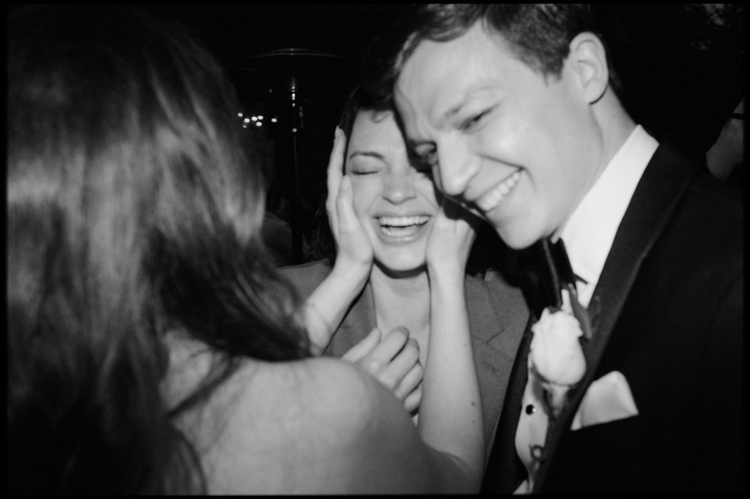 Joyful bride laughing as guest cups her face during black and white wedding celebration photo.