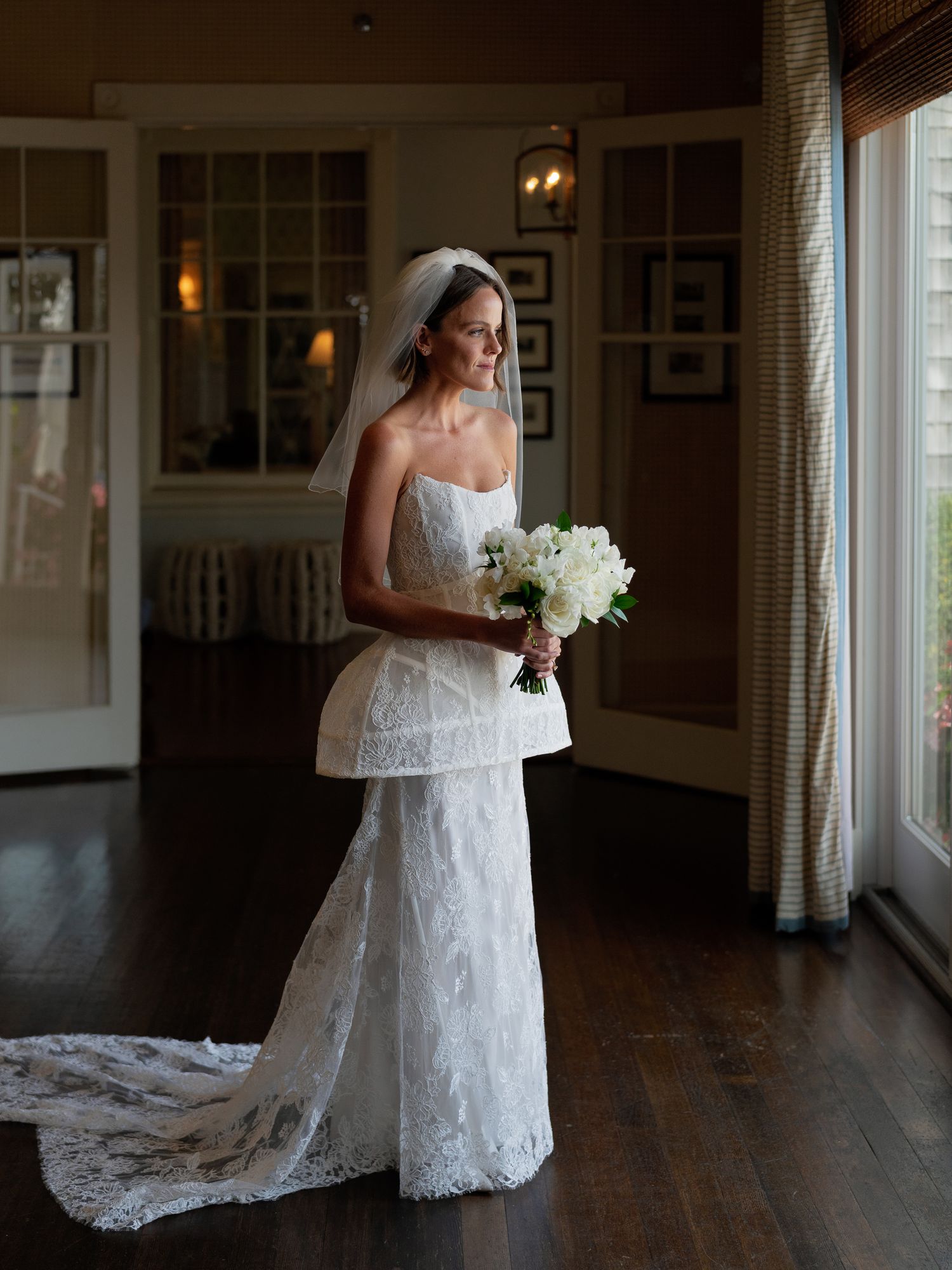 Elegant bride in strapless lace wedding gown with peplum and veil holding white bouquet indoors.