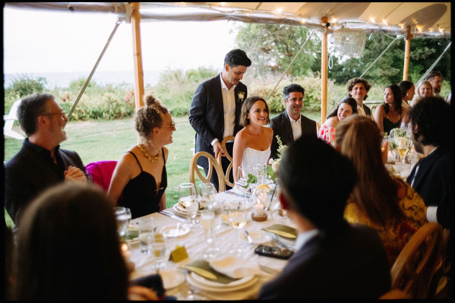 Bride and groom greet guests at decorated table during outdoor tented wedding reception.
