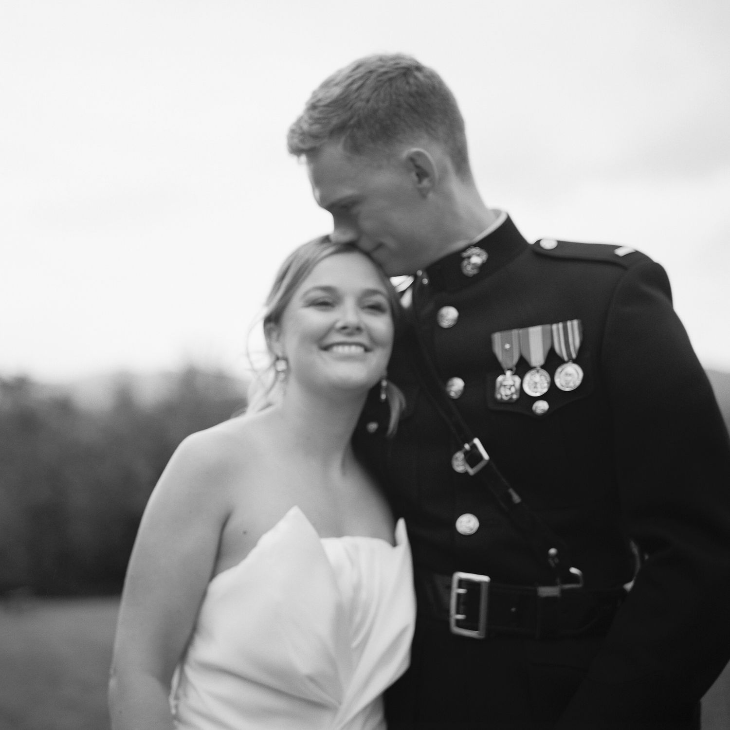 Black and white portrait of a smiling bride leaning on her Marine groom in dress uniform outdoors.