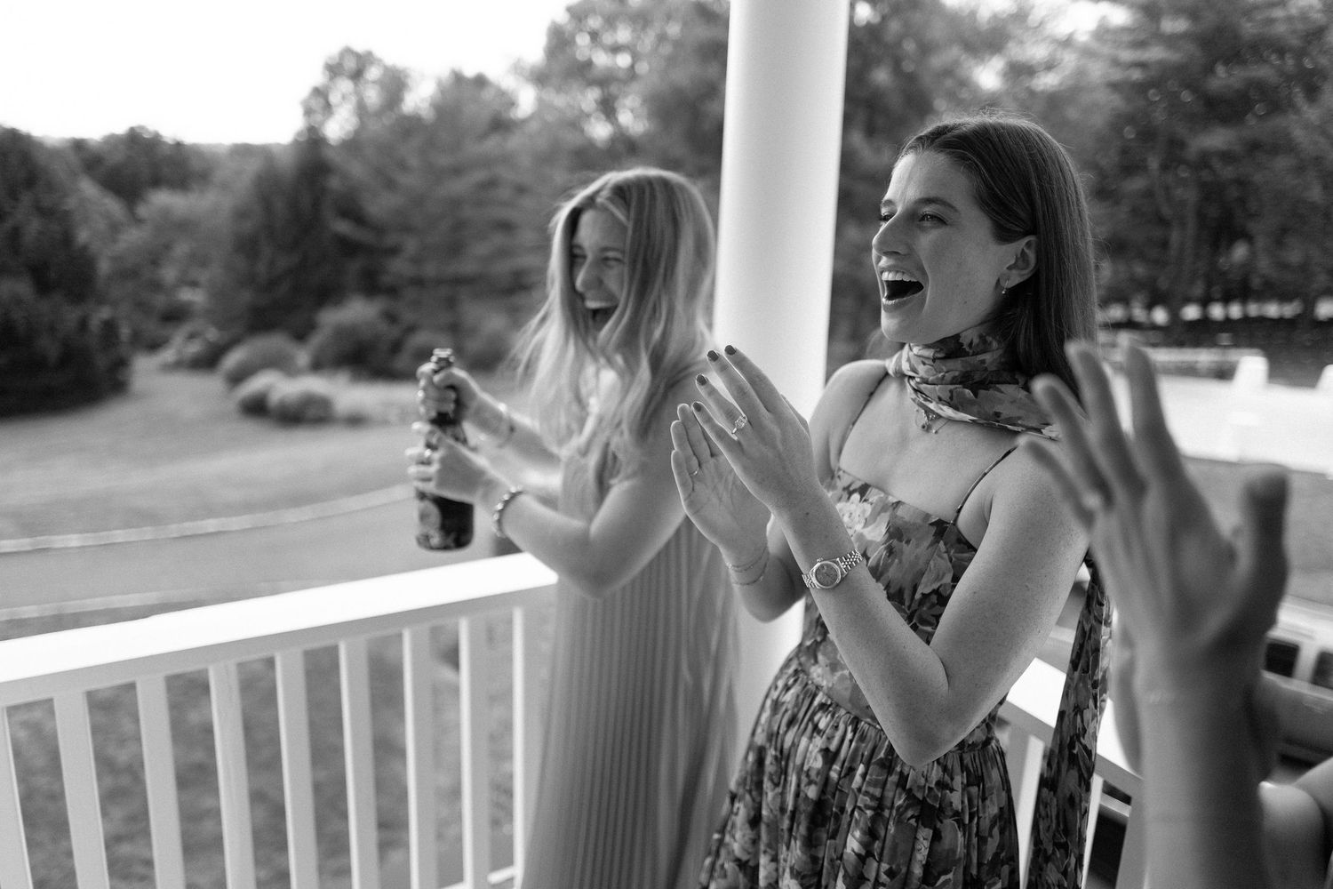 Two women laughing and celebrating with champagne on a porch at an outdoor wedding event.