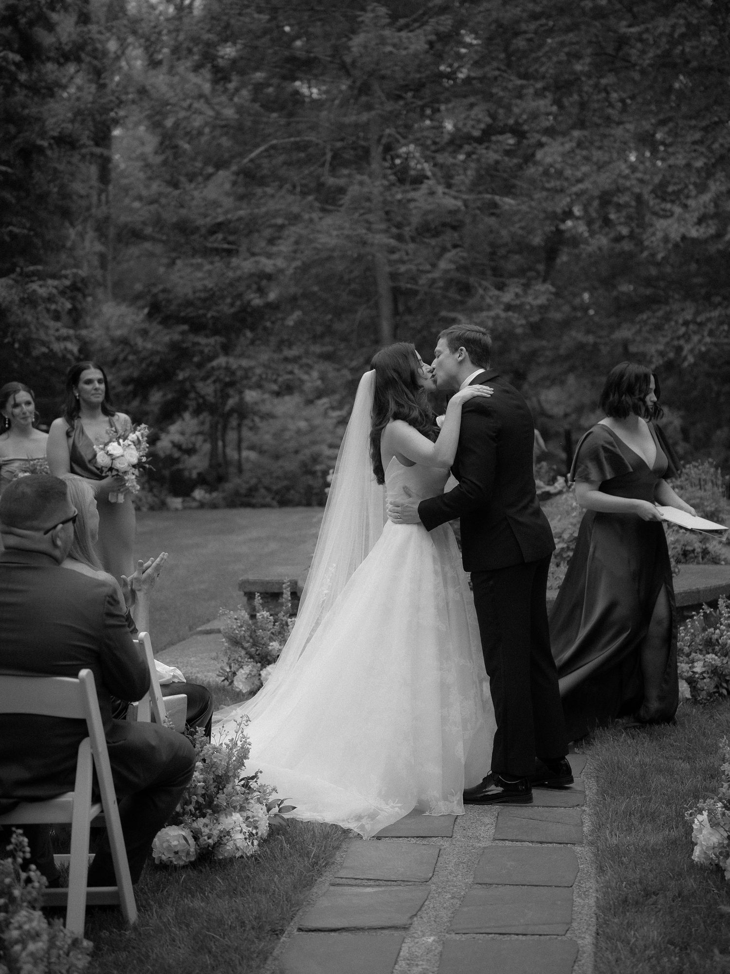 Bride and groom share their first kiss at an outdoor wedding ceremony surrounded by guests.