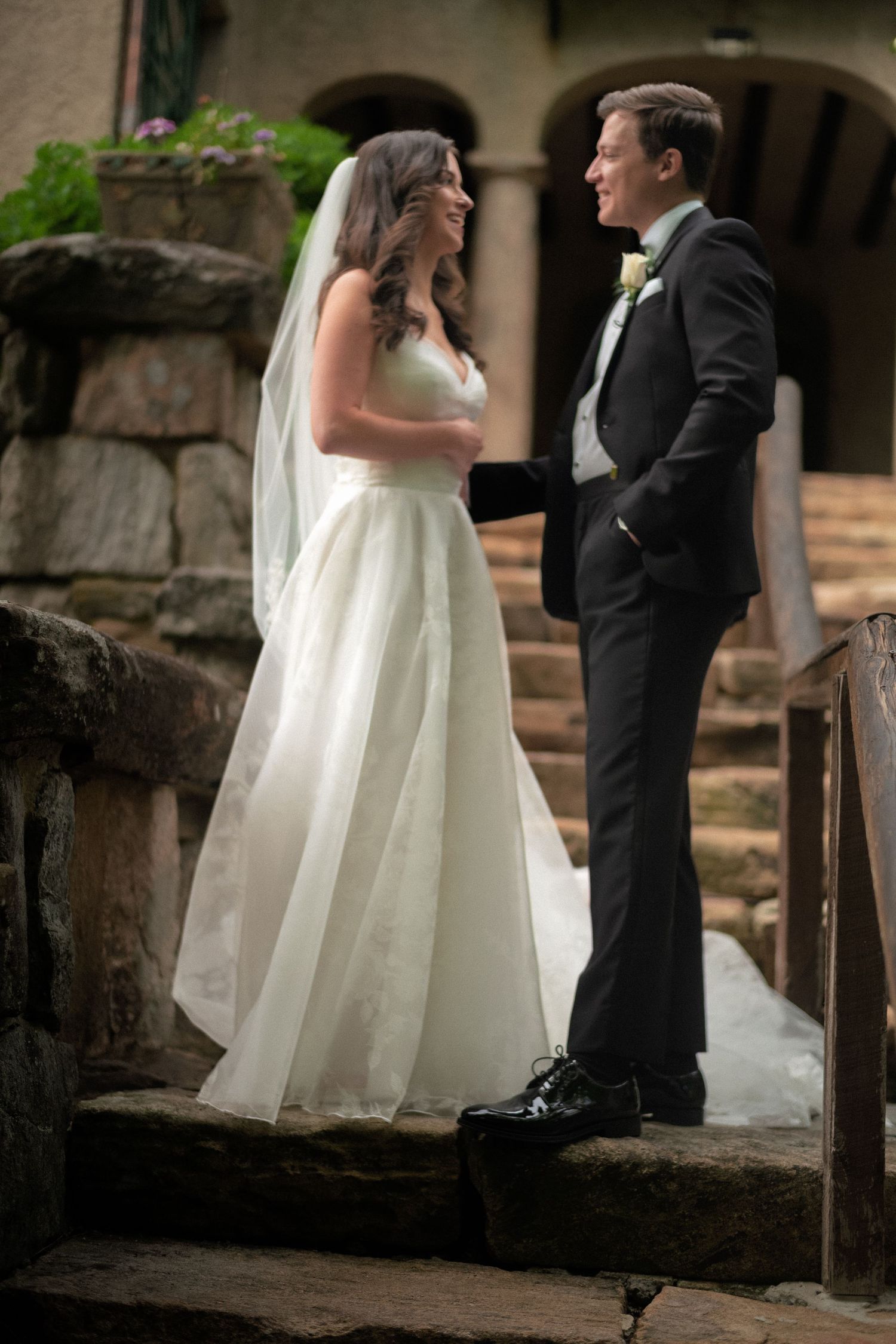 Bride in white gown and groom in black tuxedo exchange vows on stone steps at wedding venue.