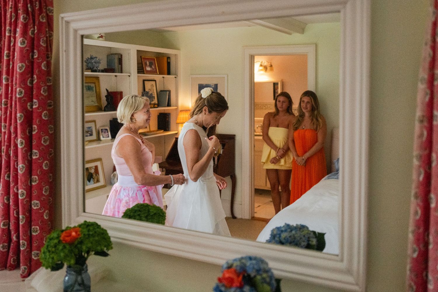 Mirror reflection captures bride getting ready with bridesmaids in colorful dresses in decorated room.