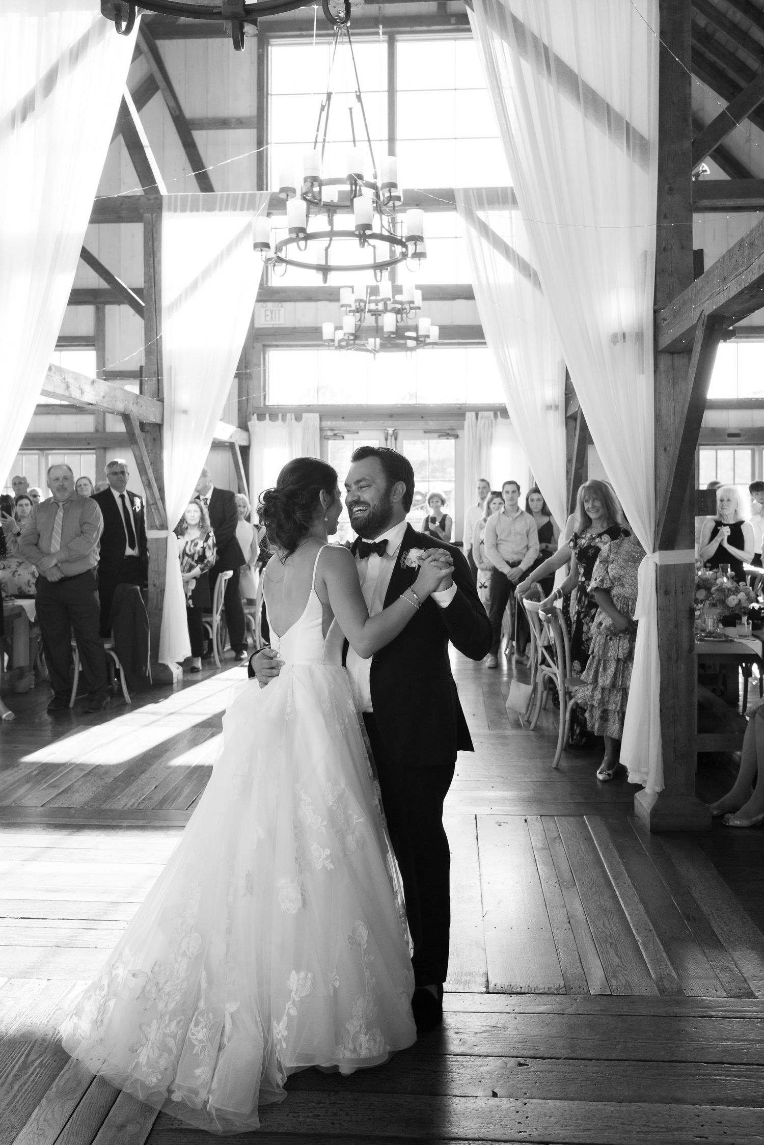 Bride and groom share their first dance in a stunning barn venue with draped white curtains and chandelier.