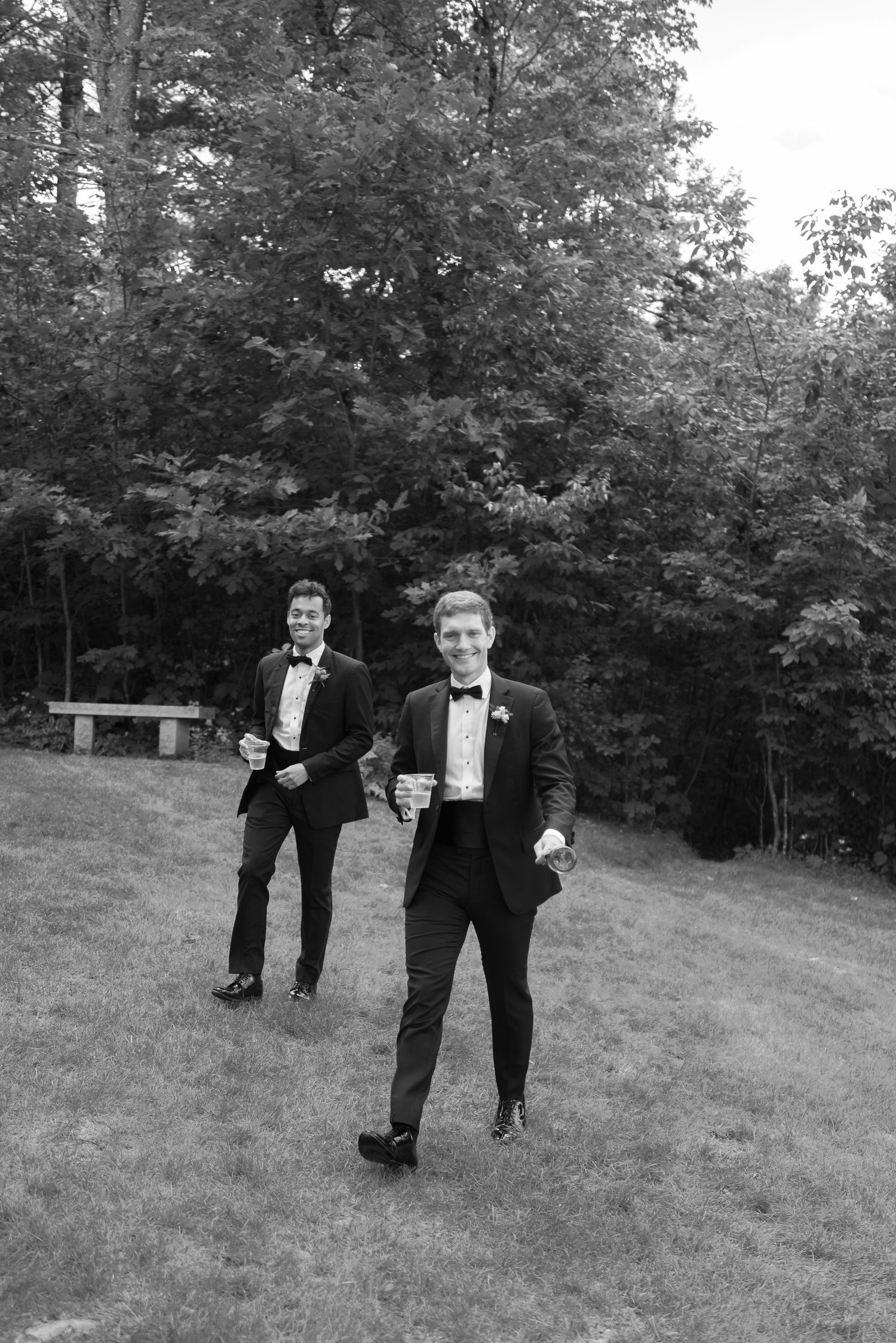 Two groomsmen in tuxedos holding drinks on grass in black and white.