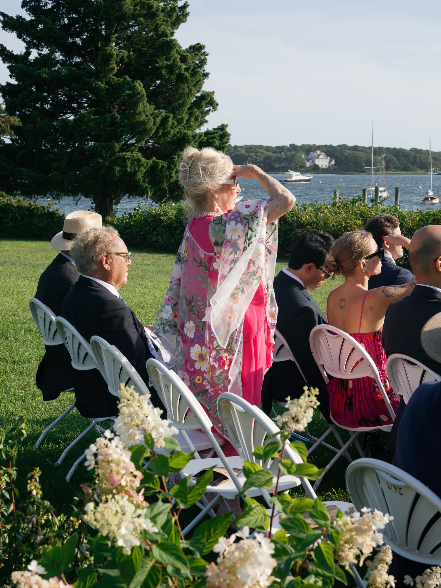 Wedding guest in floral dress shielding eyes while watching outdoor waterfront wedding ceremony on sunny day.