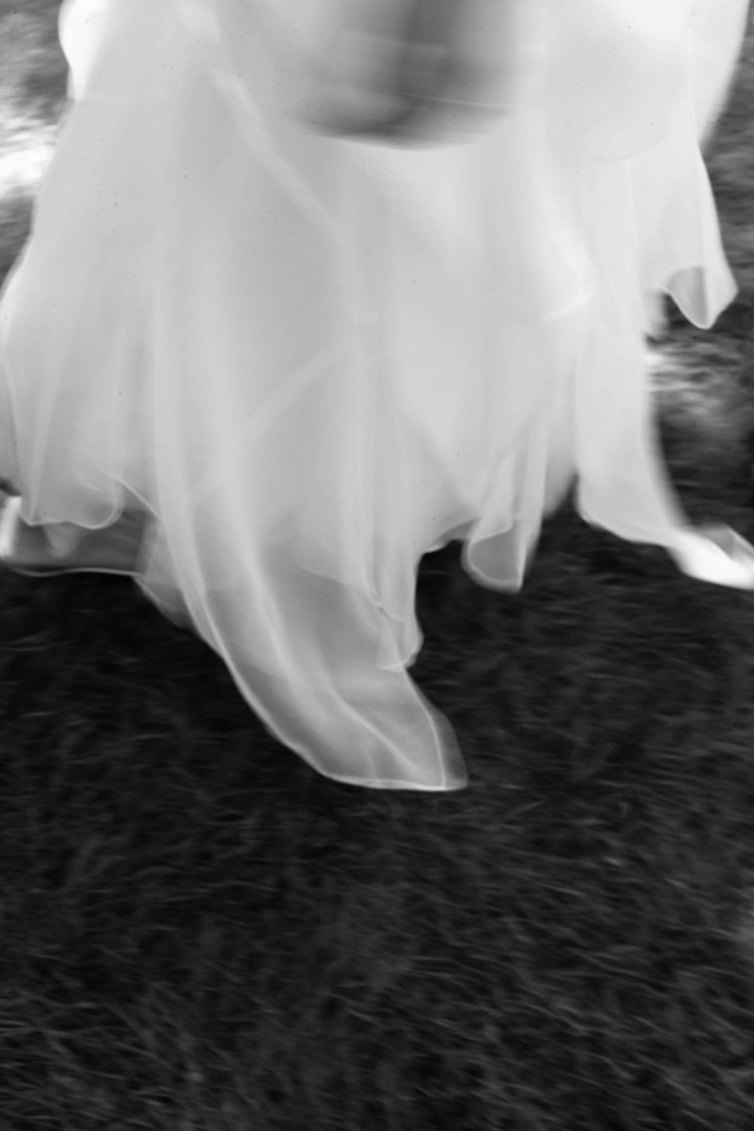 Black and white motion-blurred close-up of a flowing white wedding dress skirt and heels on grass.