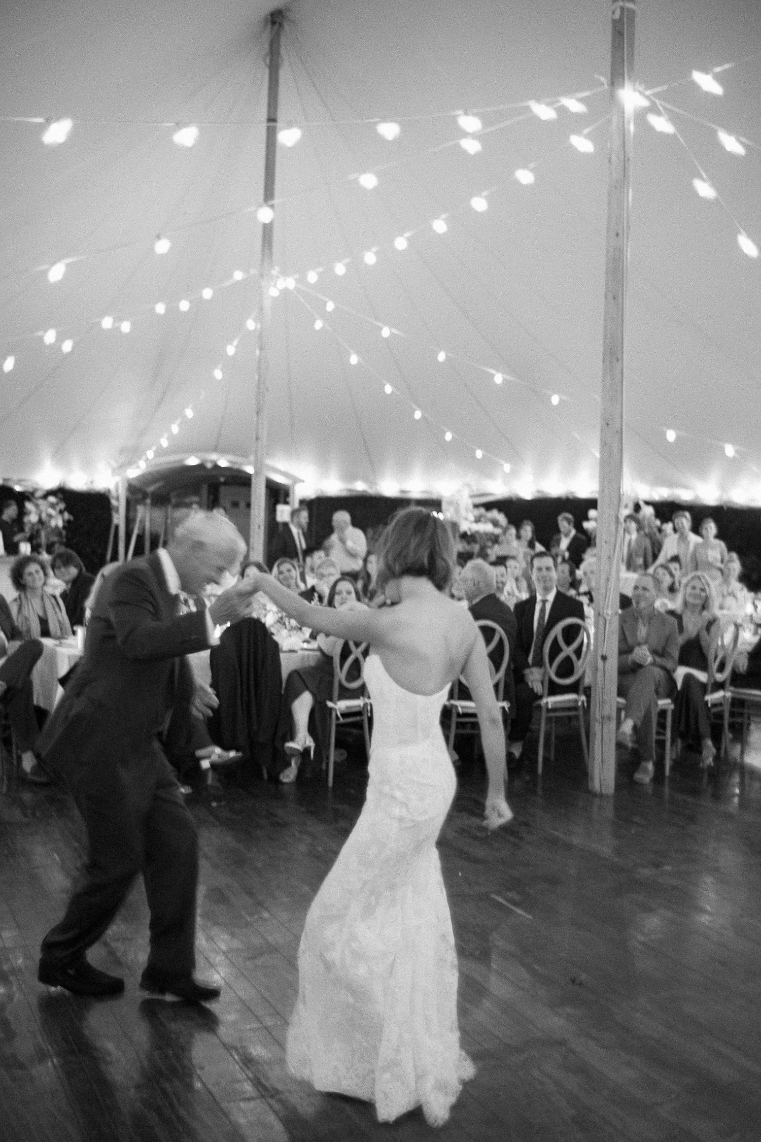 Black and white photo of bride and groom sharing first dance under string lights in elegant wedding tent.