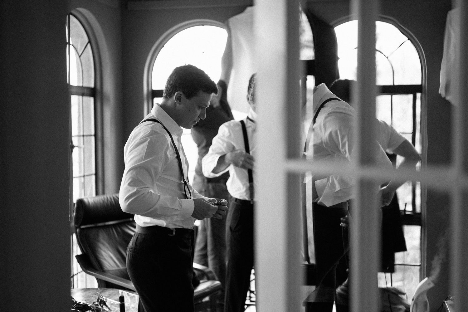 Black and white photo of a groom adjusting his suspenders while getting ready indoors near arched windows.