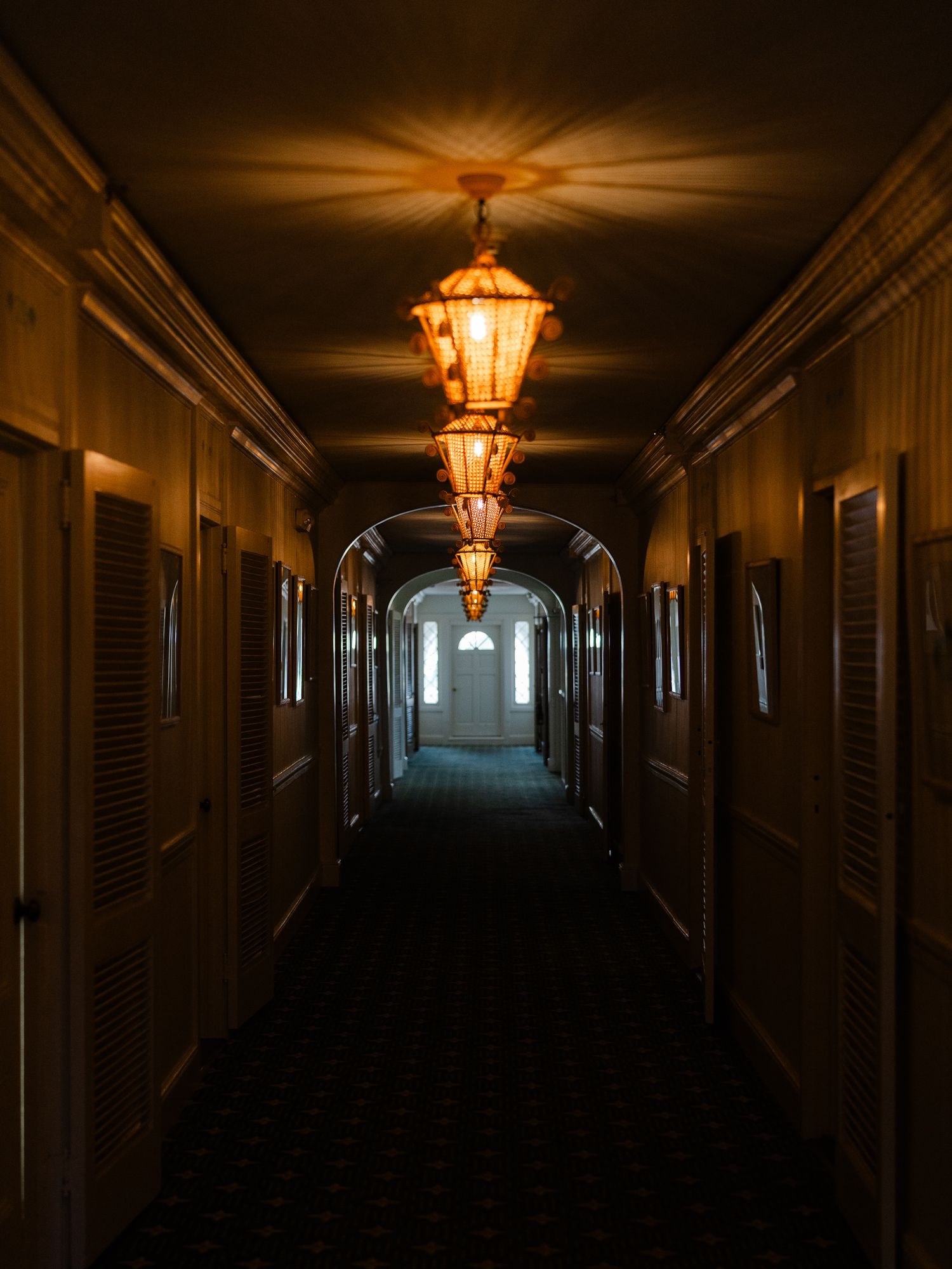 Dimly lit hotel corridor with ornate hanging lanterns casting warm golden light along an arched hallway.