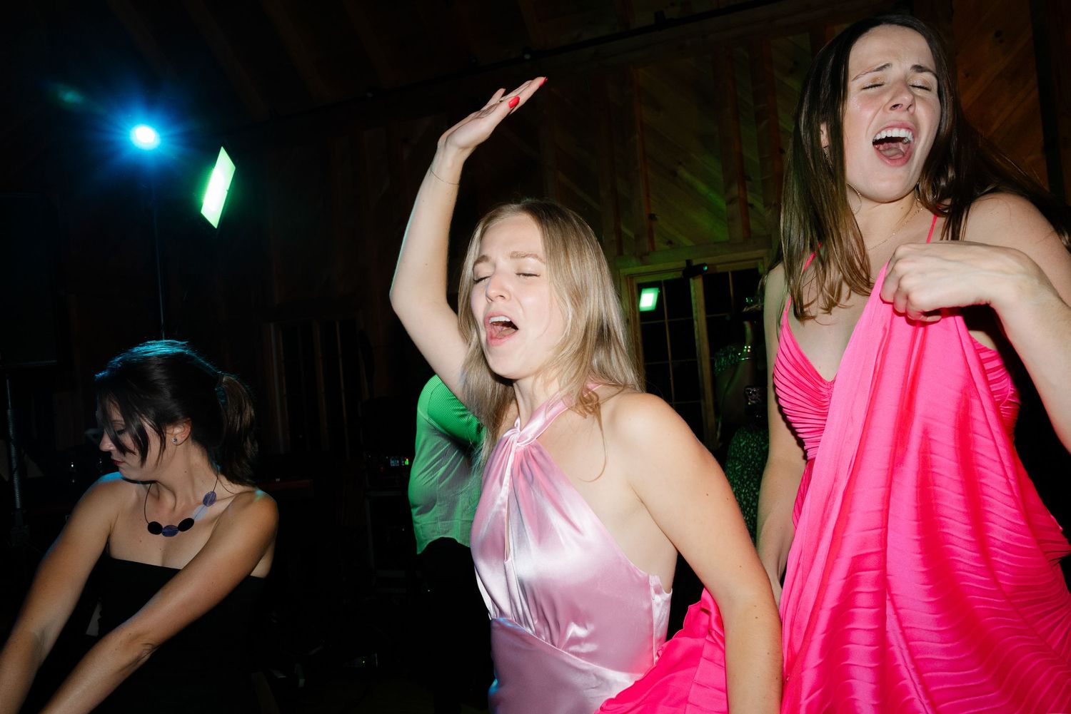 Two women dancing energetically at a wedding reception, wearing pink satin dresses under colorful party lights.