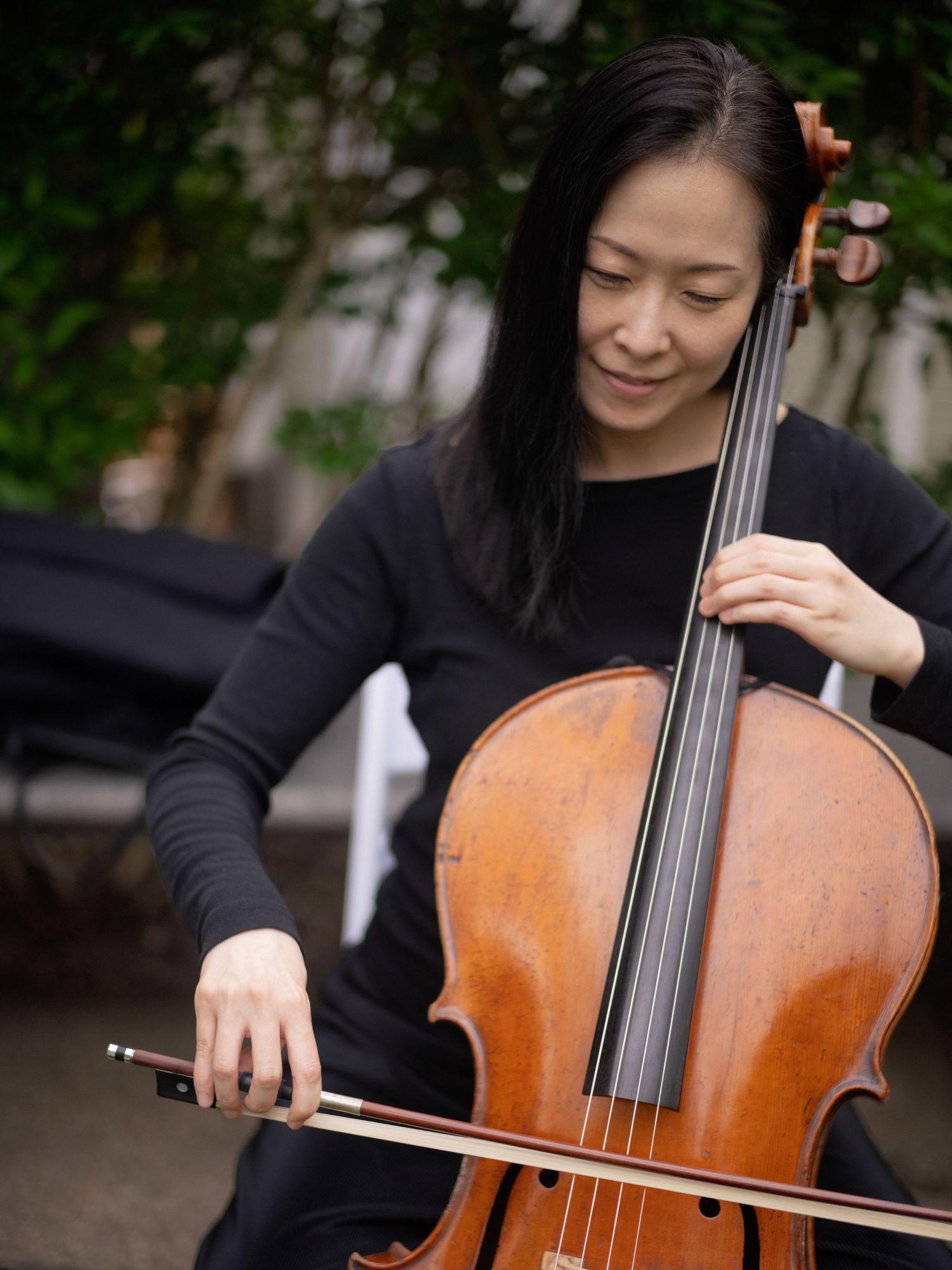 A cellist in black attire plays her wooden cello outdoors during an elegant wedding ceremony.