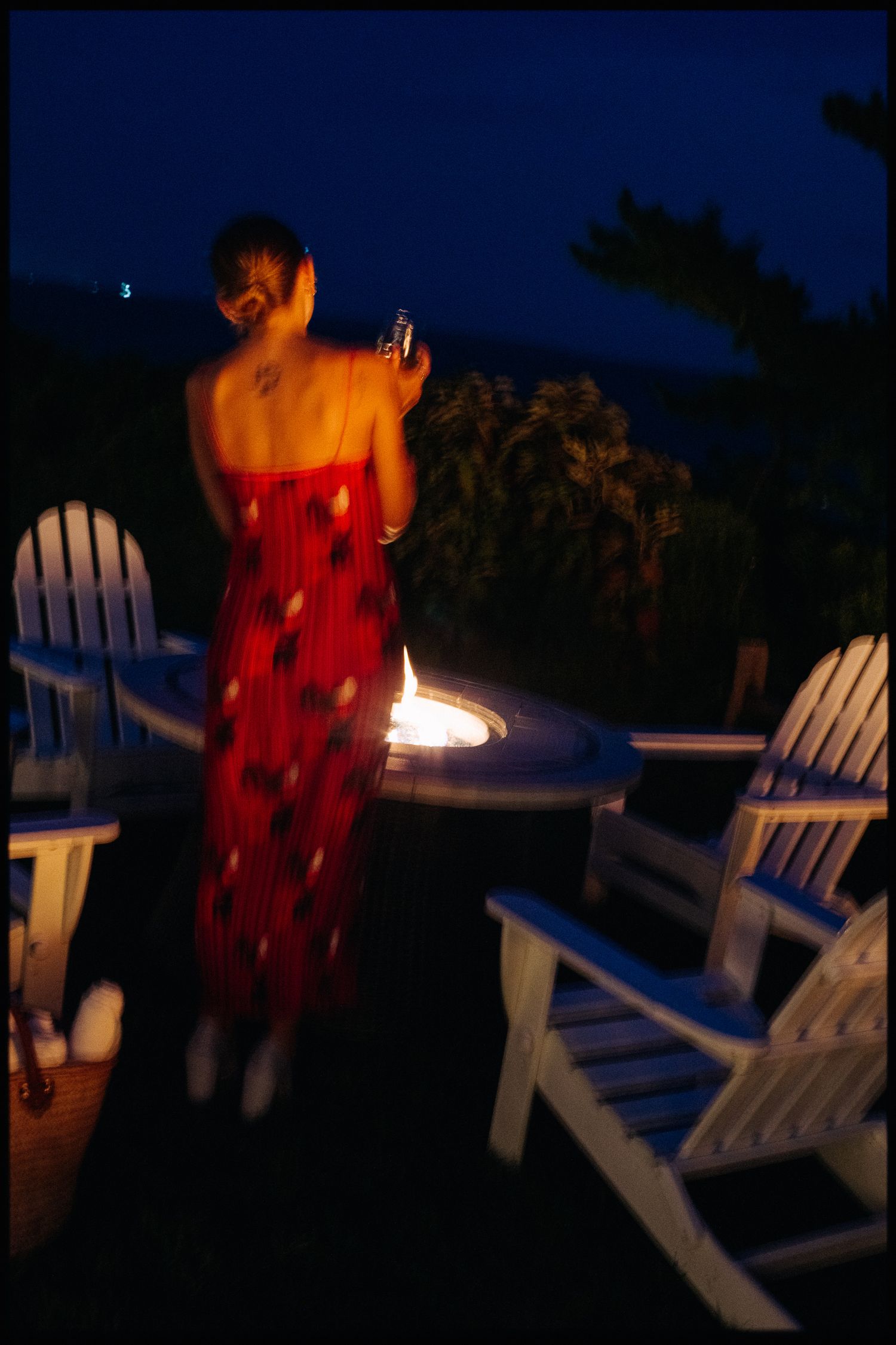 Woman in red floral dress standing near a fire pit surrounded by white Adirondack chairs at night.