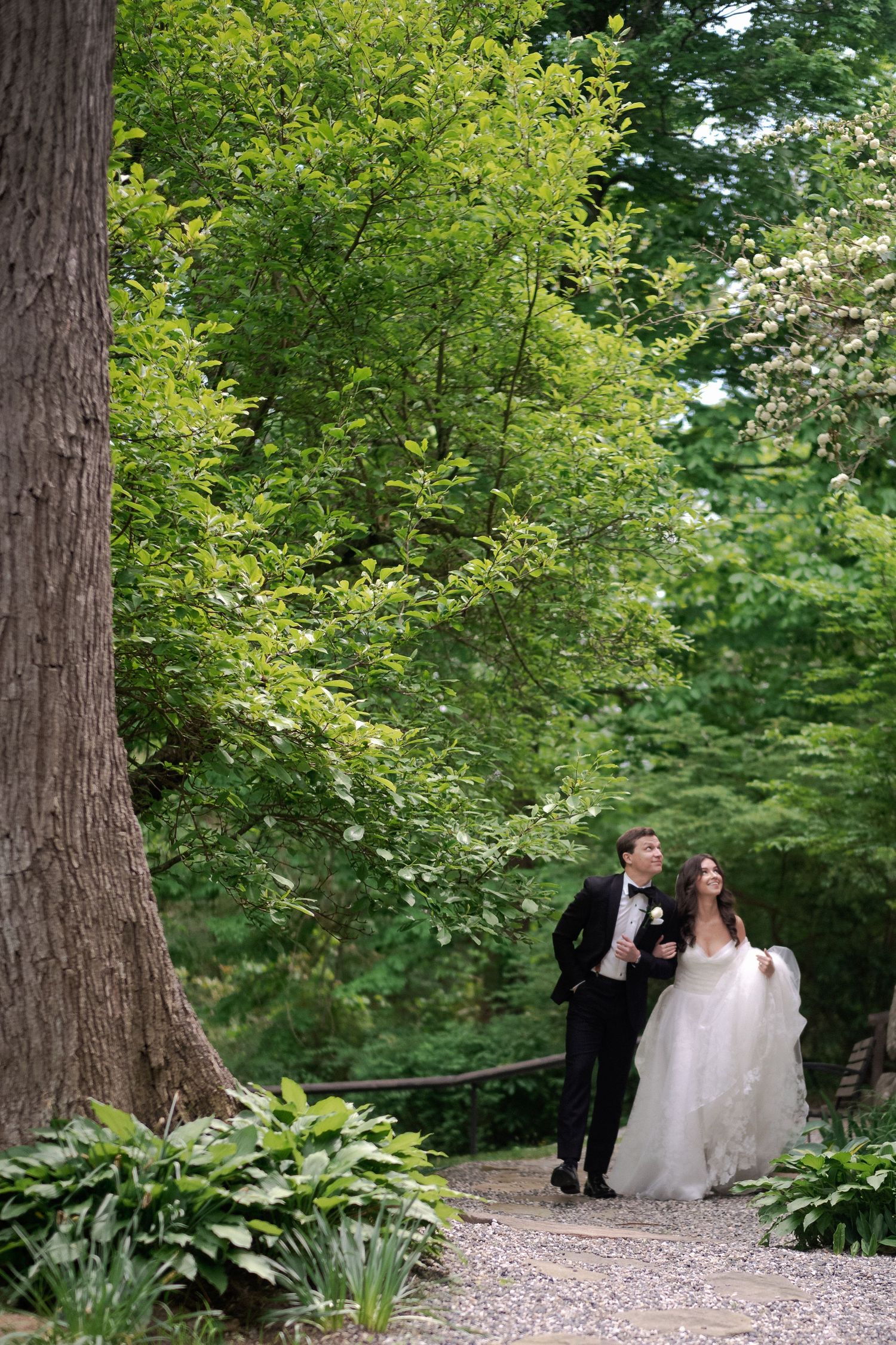 Bride and groom walk hand in hand along a lush green garden path on their wedding day.
